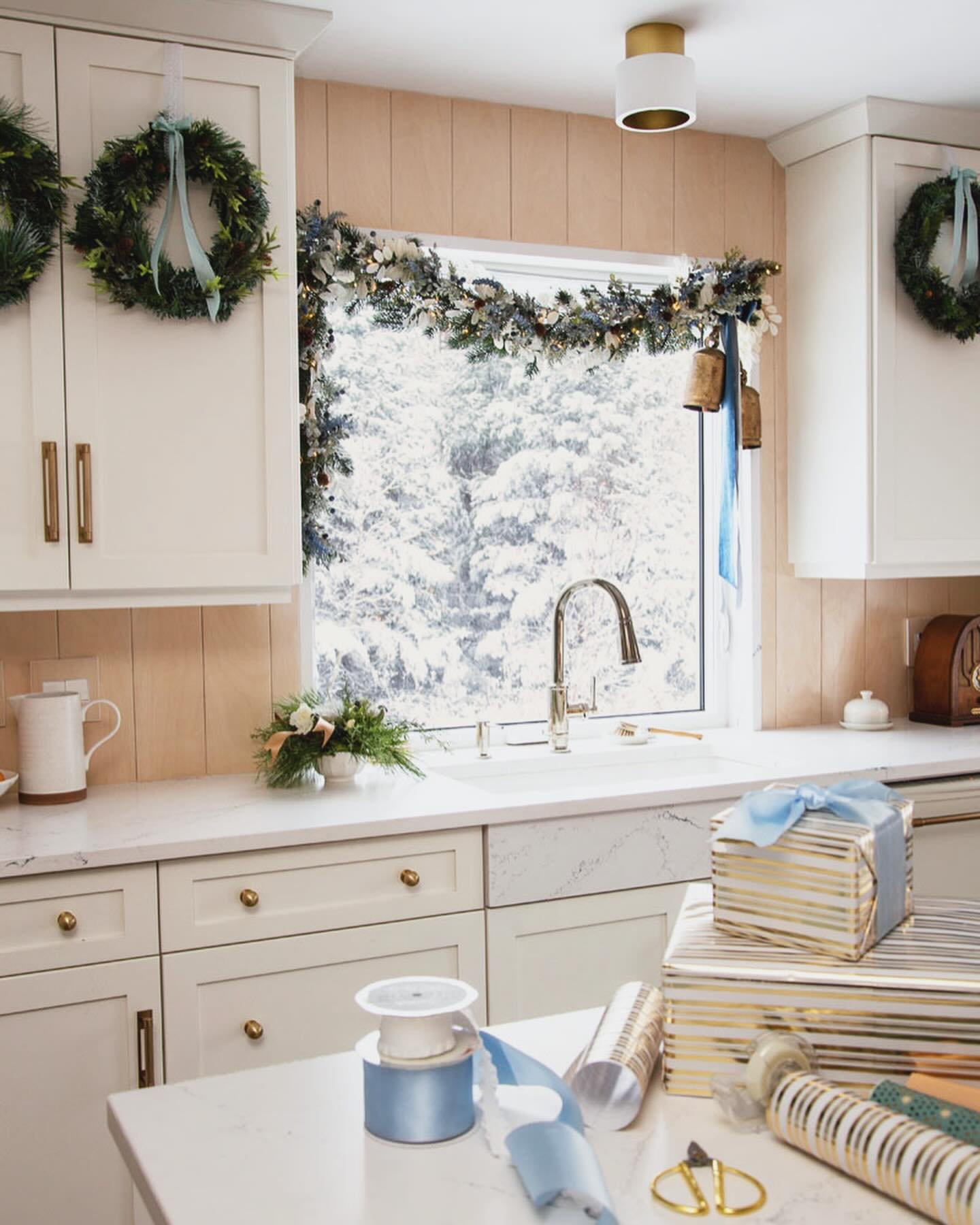 A contemporary kitchen decorated with a pastel winter theme, featuring creamy white cabinets, gold hardware, and a window framed by delicate garland with soft blue accents, large gold bells, and coordinating blue ribbon.