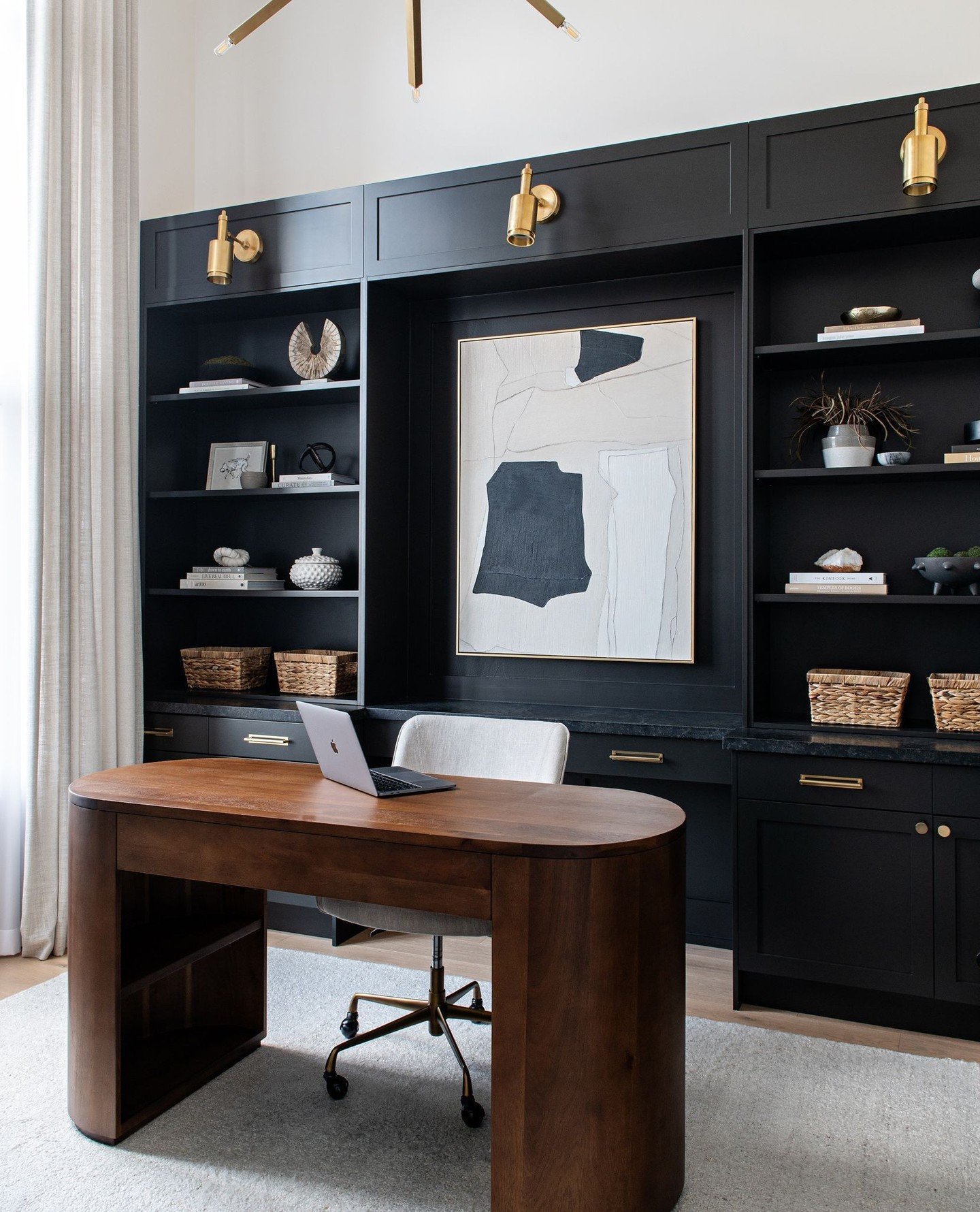 Modern home office featuring a matte black built-in wall unit with brass sconces, a light gray rug, and a dark walnut oval-shaped desk.