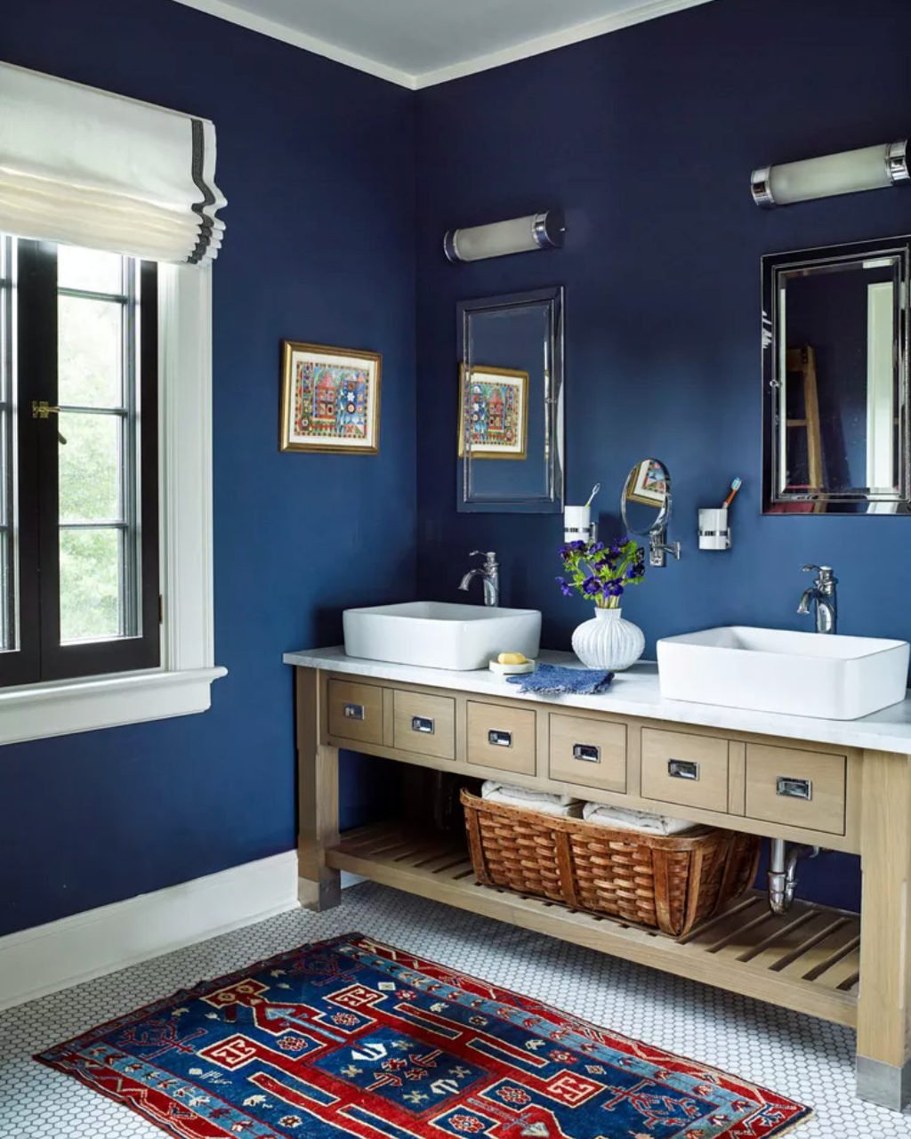 A navy blue bathroom featuring a light wood double vanity, white hexagonal tile floor, and a small, richly patterned red and blue Persian-style rug.