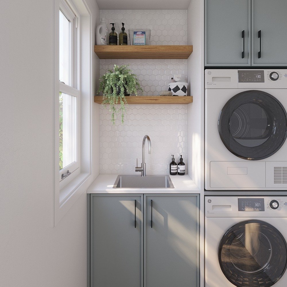 A small, bright laundry room featuring a stainless steel sink, sage-green lower cabinets, white hexagon tile backsplash, wooden floating shelves, and a stacked washer and dryer unit.