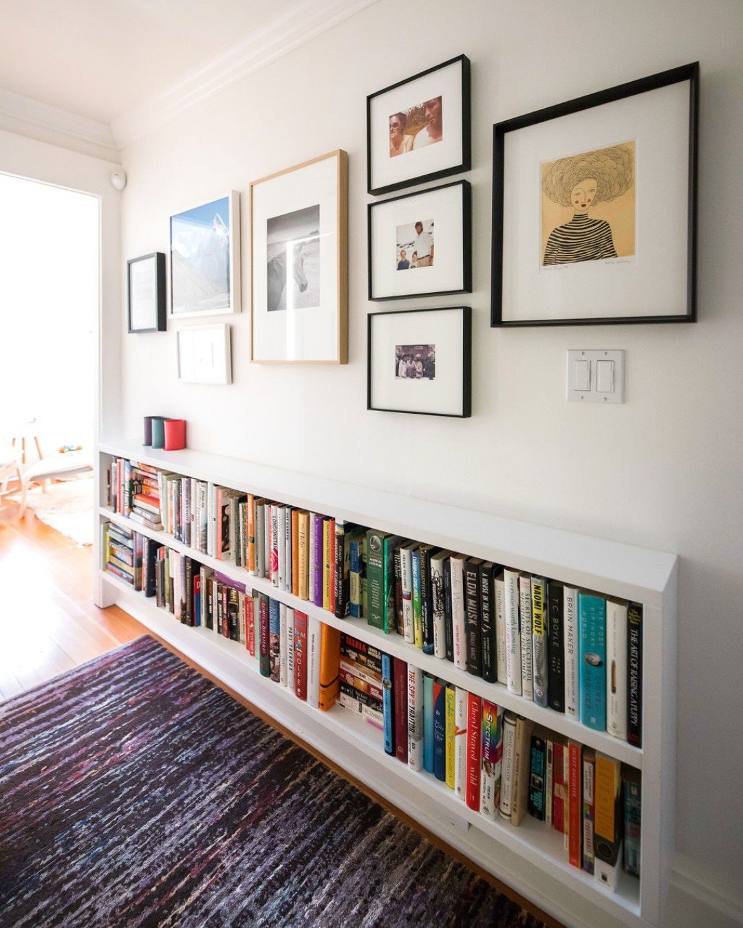 A long, low white recessed shelf built into a narrow hallway wall is filled with a colorful collection of books and topped with a gallery wall of framed art.
