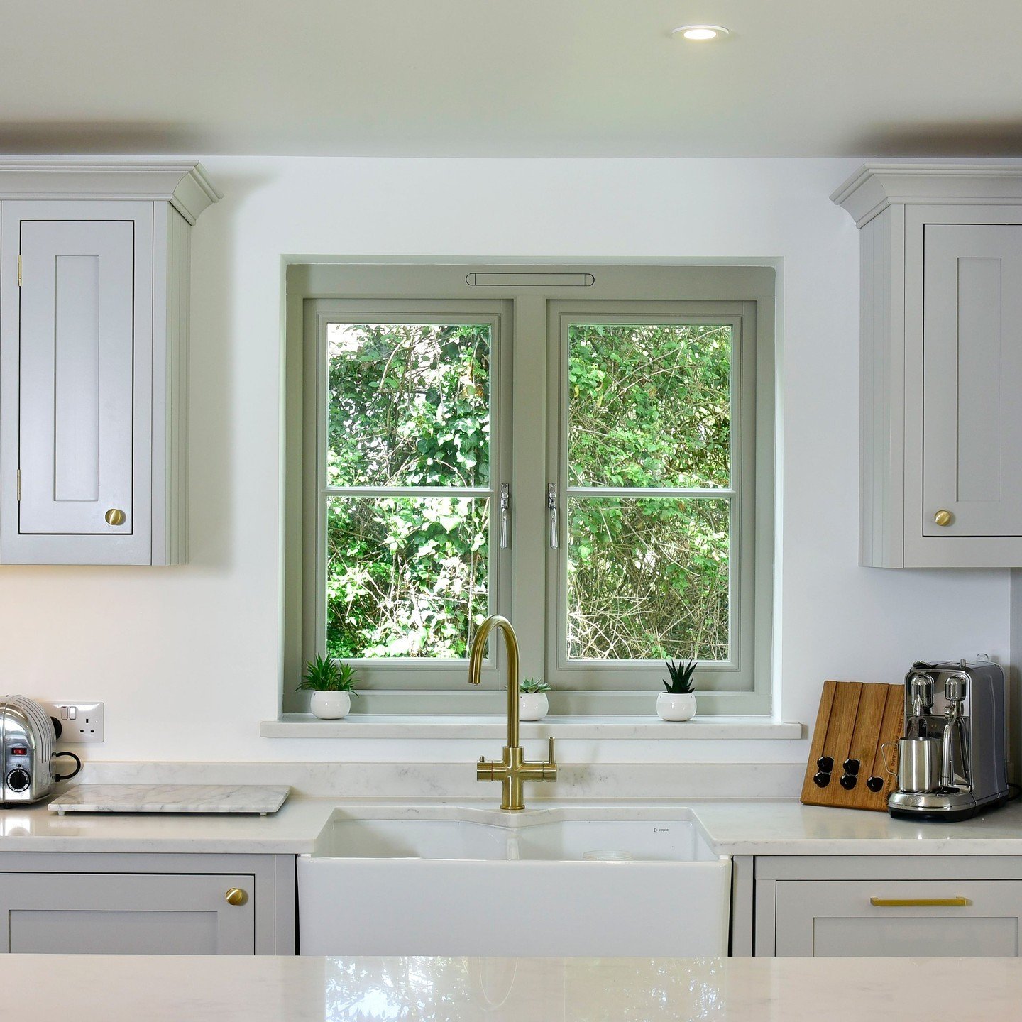 Traditional kitchen with grey shaker cabinets. A brass faucet is centered on a white farmhouse sink below a sage green sash window looking out to lush green foliage.