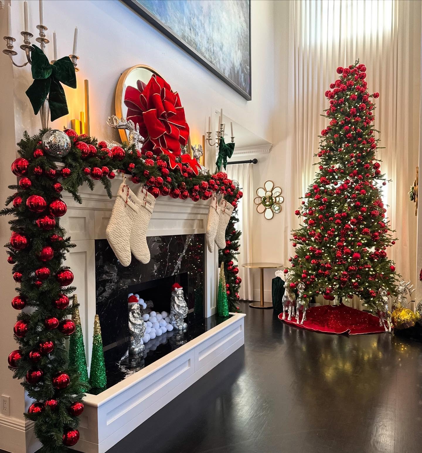White mantel decorated with thick evergreen garland, large red ornaments, white stockings, and a huge red velvet bow.