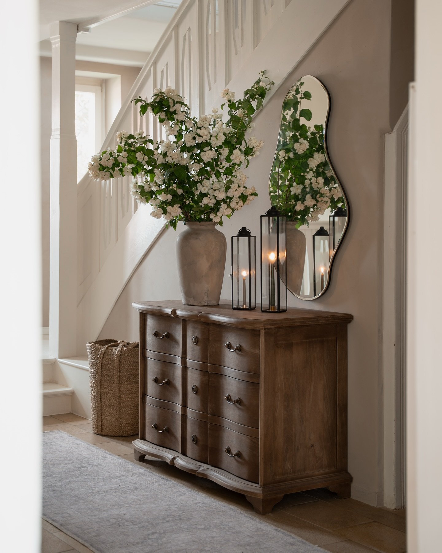A rustic wood console table topped with a tall vase of white flowers a curved black mirror and black lanterns.