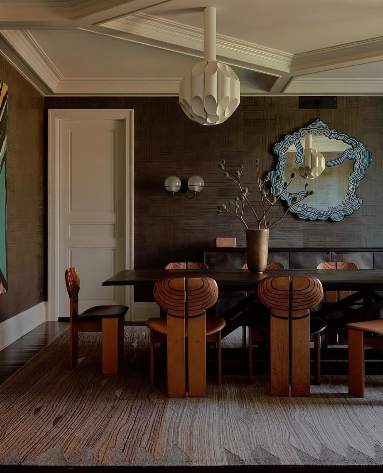 A moody dining room featuring textured dark brown walls, unique wooden chairs, a long dark table, and a sculptural white pendant light.