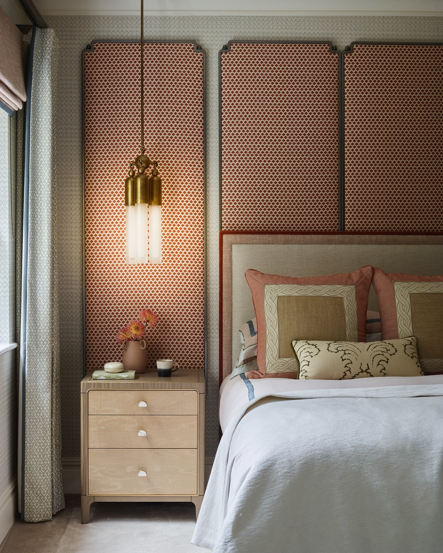 Floor-to-ceiling upholstered headboard panels in a terracotta geometric print, separated by thin grey trim, flanked by a light wood nightstand and brass pendant light.