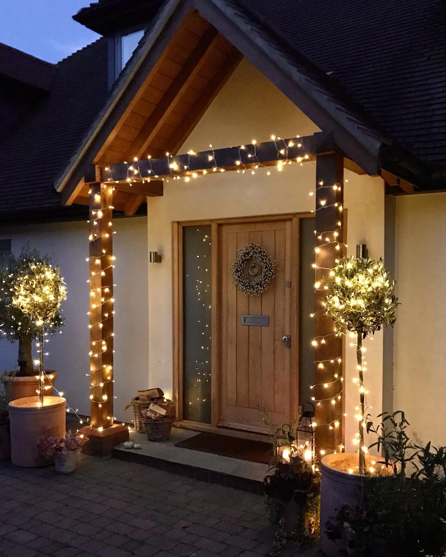 Warm white string lights wrap wooden porch columns and two potted trees flanking a light wood front door with a simple wreath.