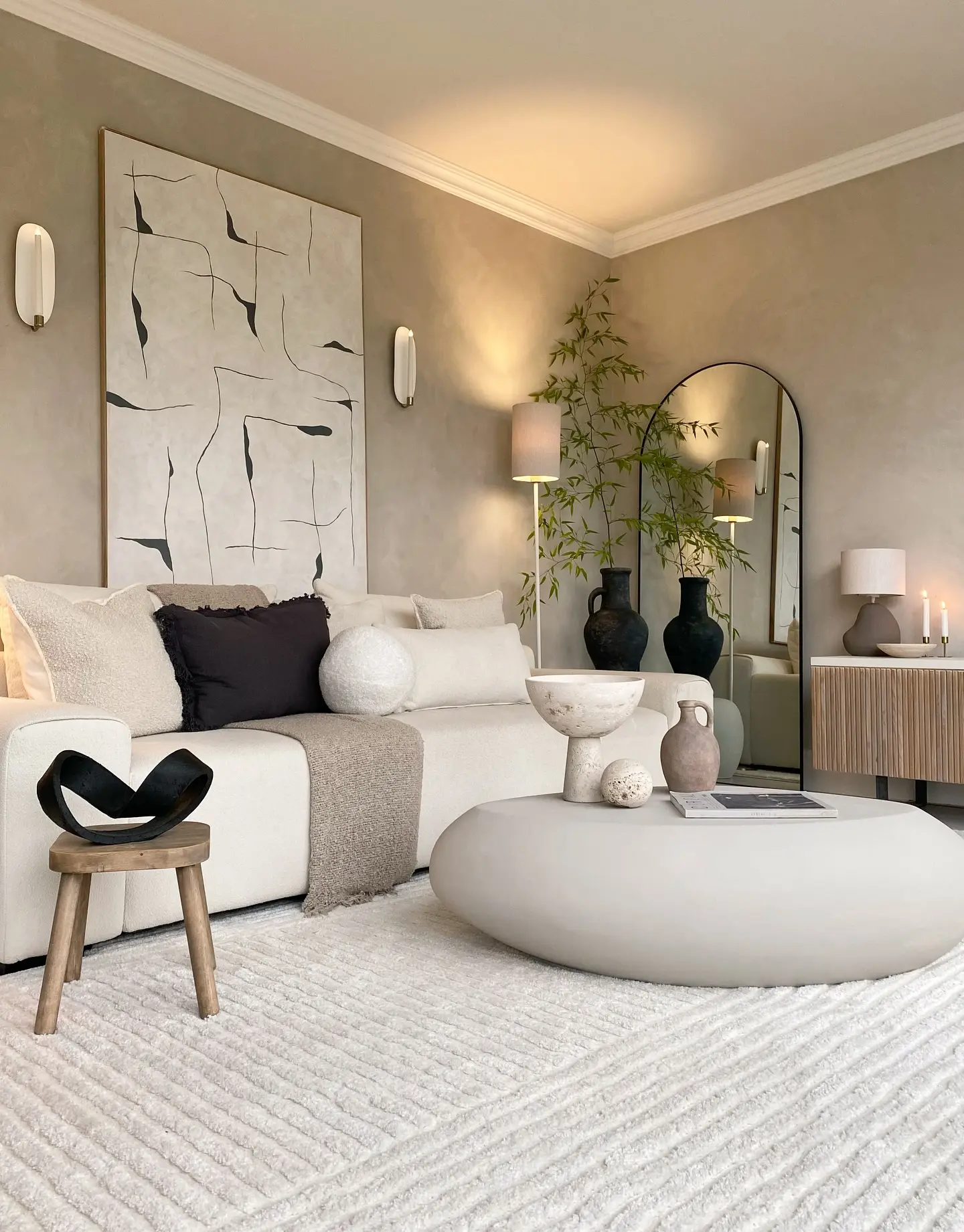 Oversized vertical abstract line art painting in black on a beige canvas, hung above a white sectional sofa in a neutral-toned living room with a rounded mirror.