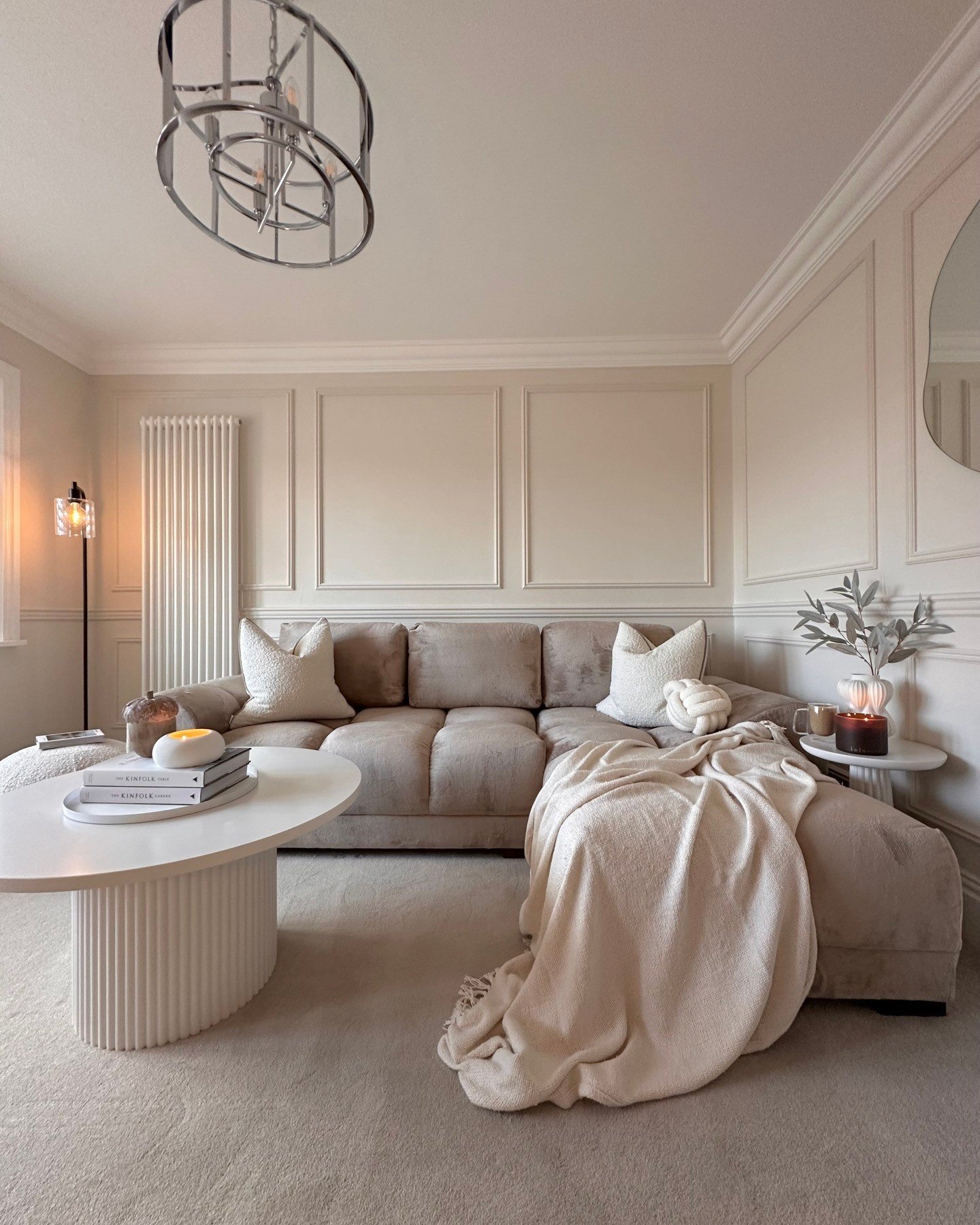 A cozy, modern beige living room featuring wainscoting, a plush velvet sectional sofa with knit throws, and a round fluted white coffee table.