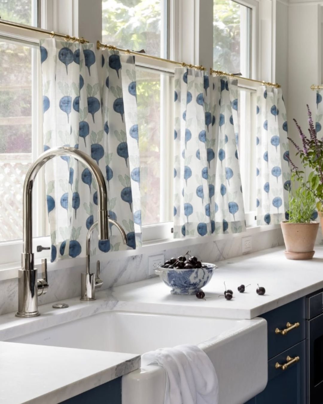 Kitchen window with white cafe curtains featuring a blue and green radish pattern hung on a brass rod over a marble countertop and farmhouse sink.
