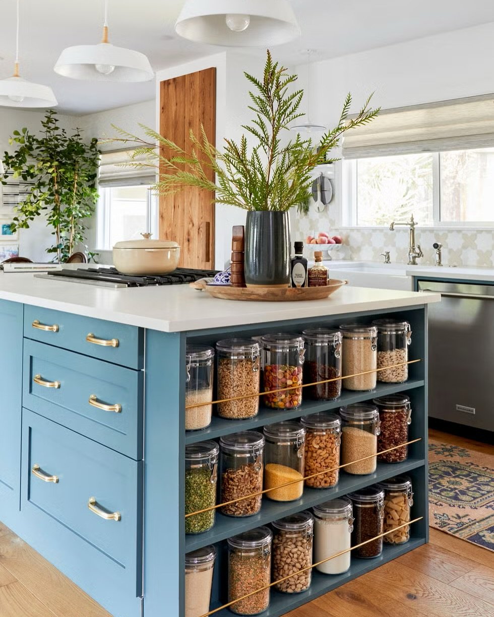 Teal kitchen island with open-side shelving displaying rows of dry goods in clear glass jars.