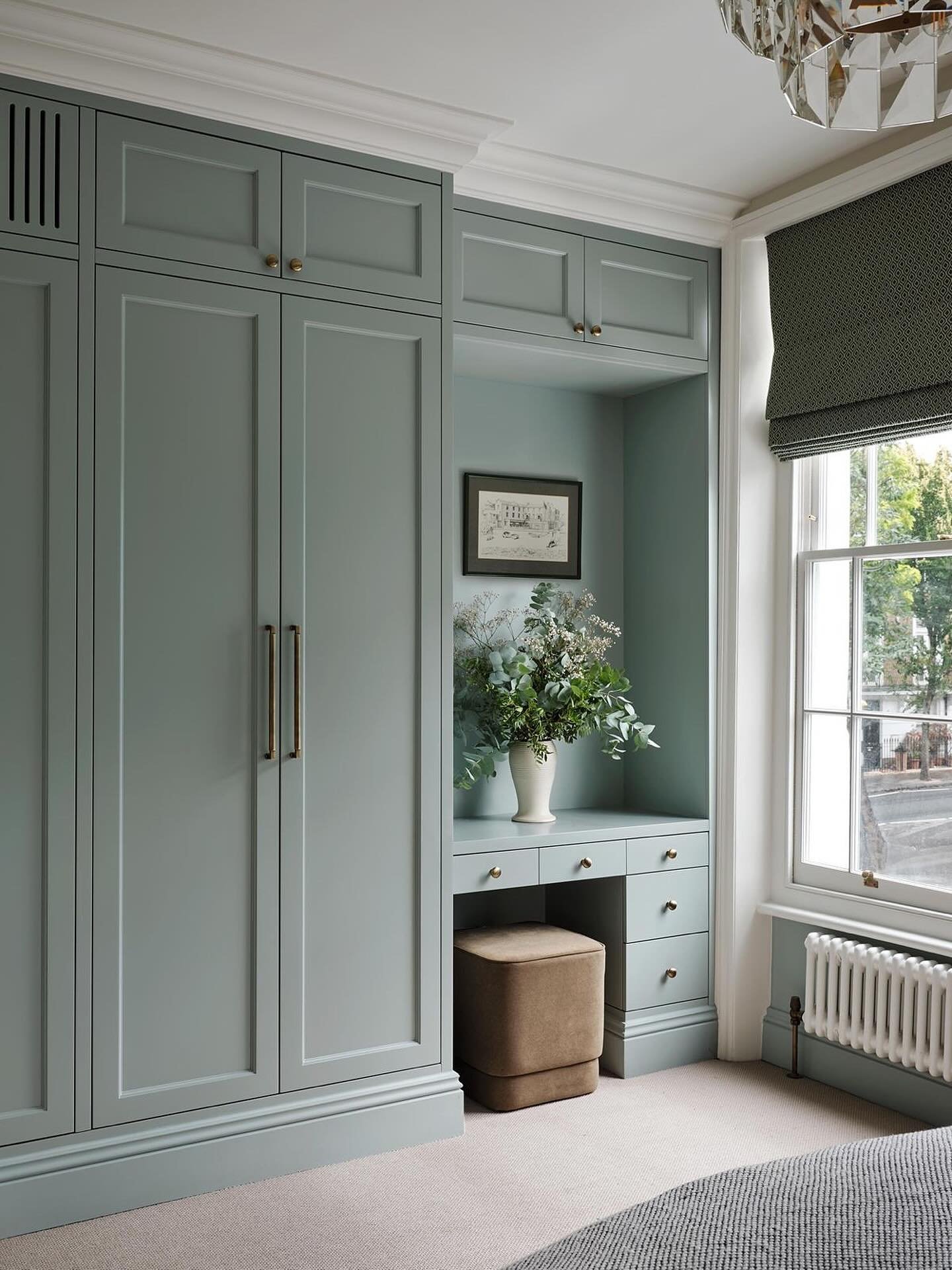 Built-in sage green shaker wardrobe with brass hardware and a recessed central vanity nook with drawers and a brown ottoman stool.