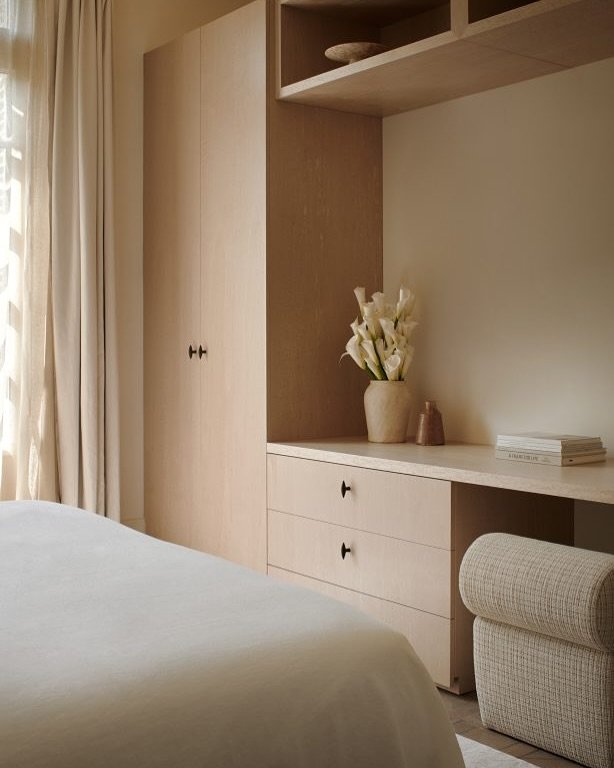 Minimalist bedroom featuring a light wood built-in unit combining a full-height wardrobe, drawers, and a desk with open shelving above.