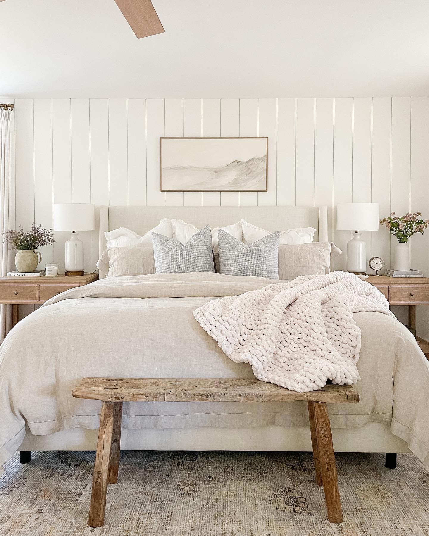 A neutral, light-filled bedroom featuring a large bed with linen bedding and a small, rustic, salvaged wood bench at the foot of the bed.