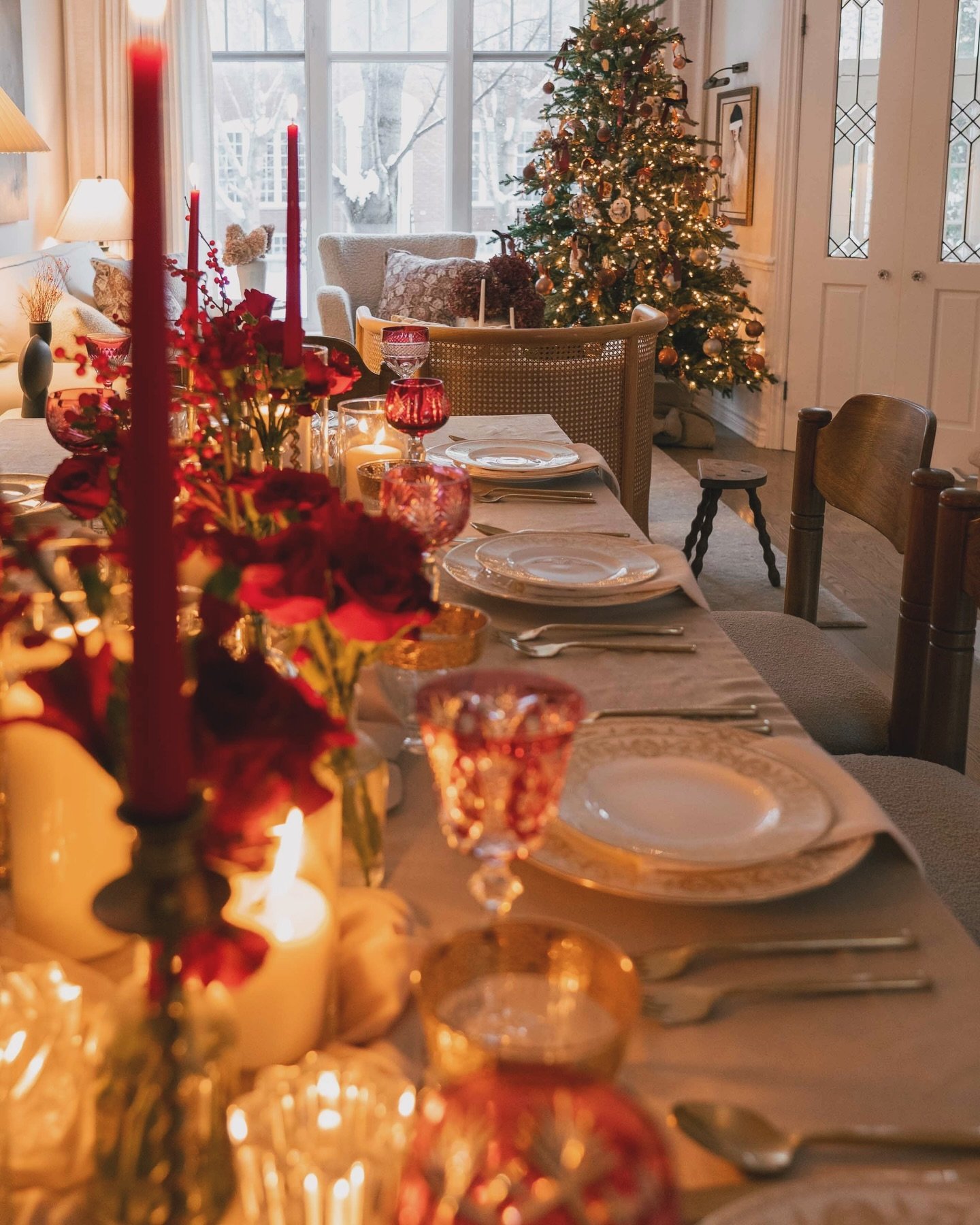 A Christmas dining table set with a beige runner, layered white and gold plates, ruby red cut glass, and dark red floral arrangements and taper candles.
