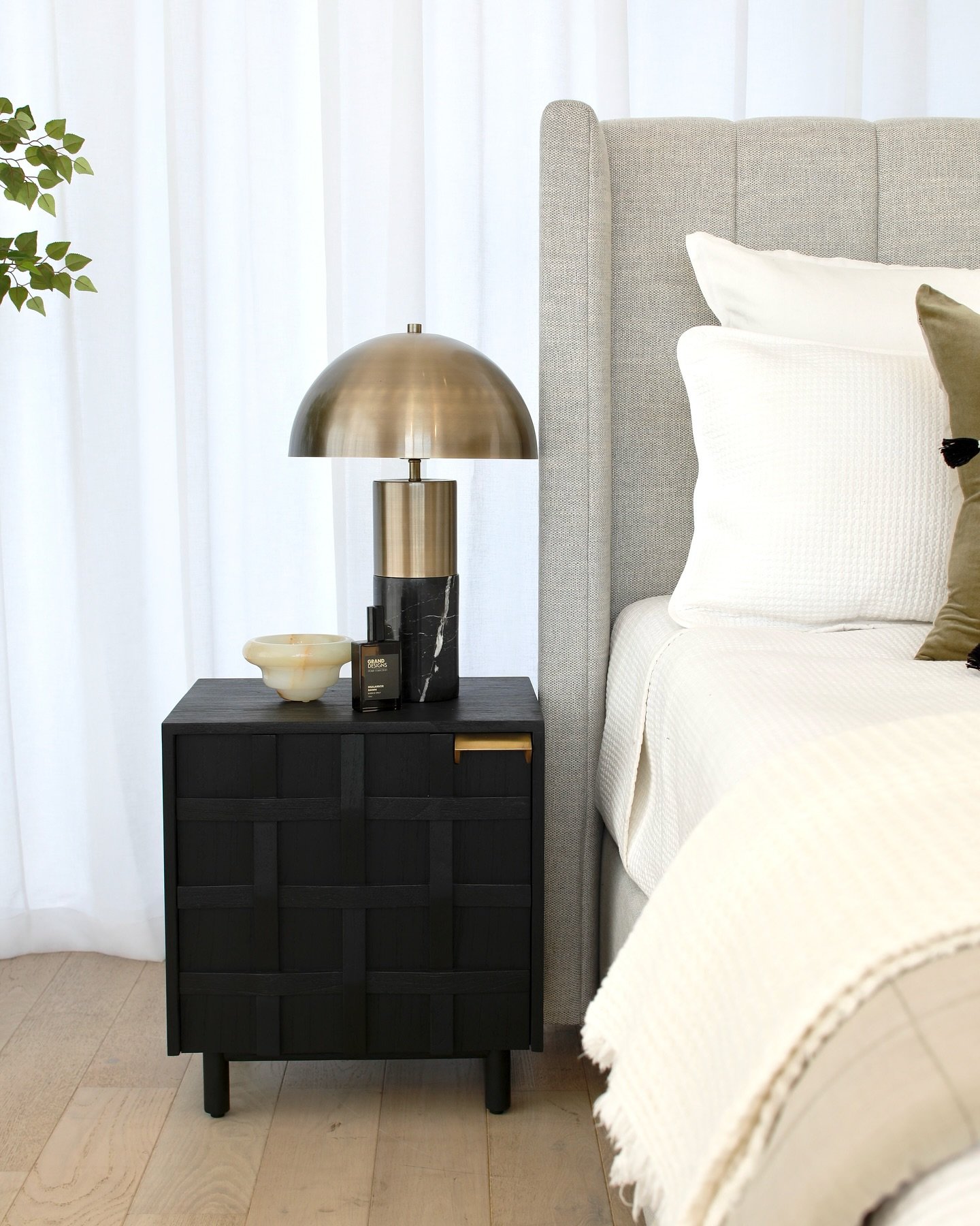 Black nightstand with woven drawer fronts next to a gray upholstered bed and brass lamp.