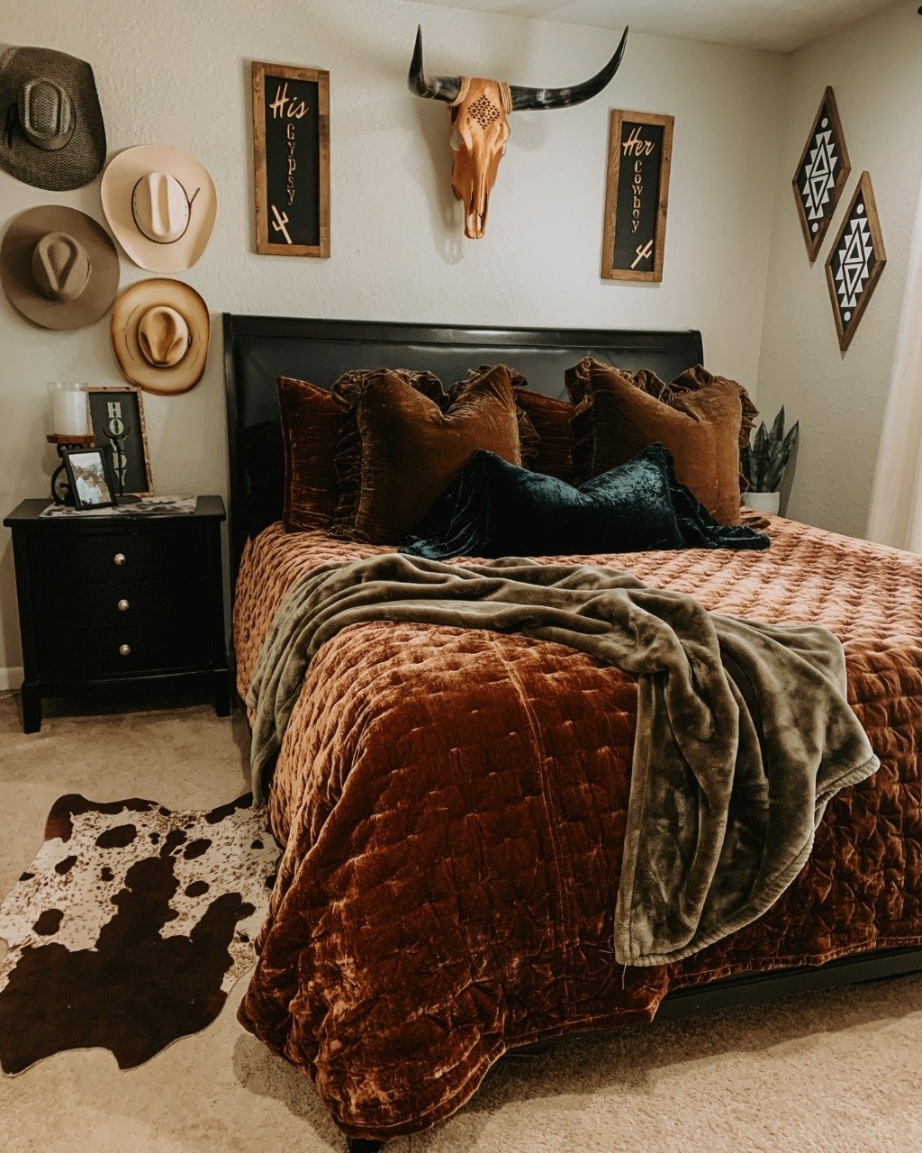 A cozy Western-themed bedroom featuring a burnt orange velvet quilt, multiple cowboy hats hung on the wall, and a cowhide rug.