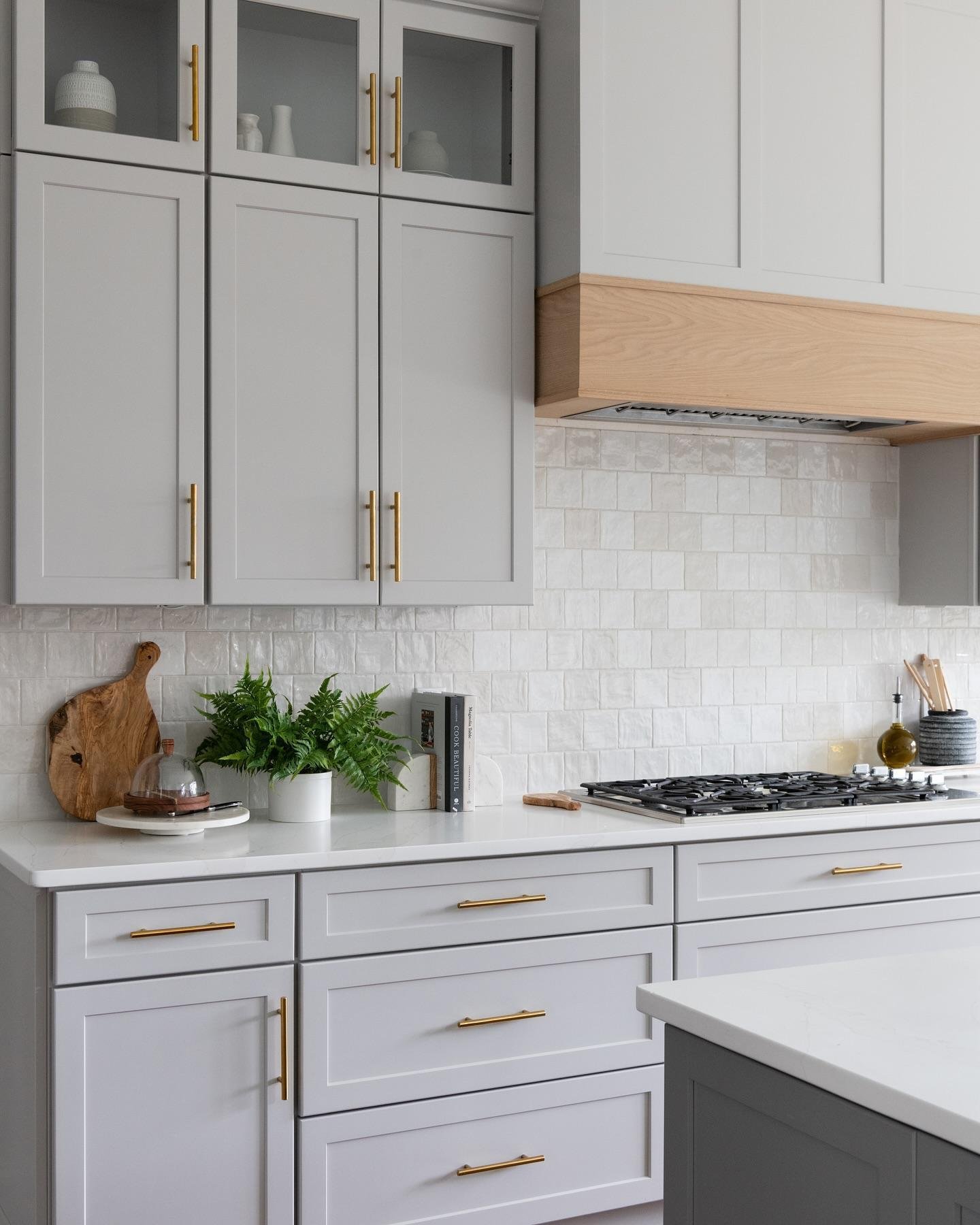 Modern kitchen with light gray cabinets, gold hardware, a natural wood range hood, and a textured, handmade-style white square tile backsplash over a white quartz countertop.