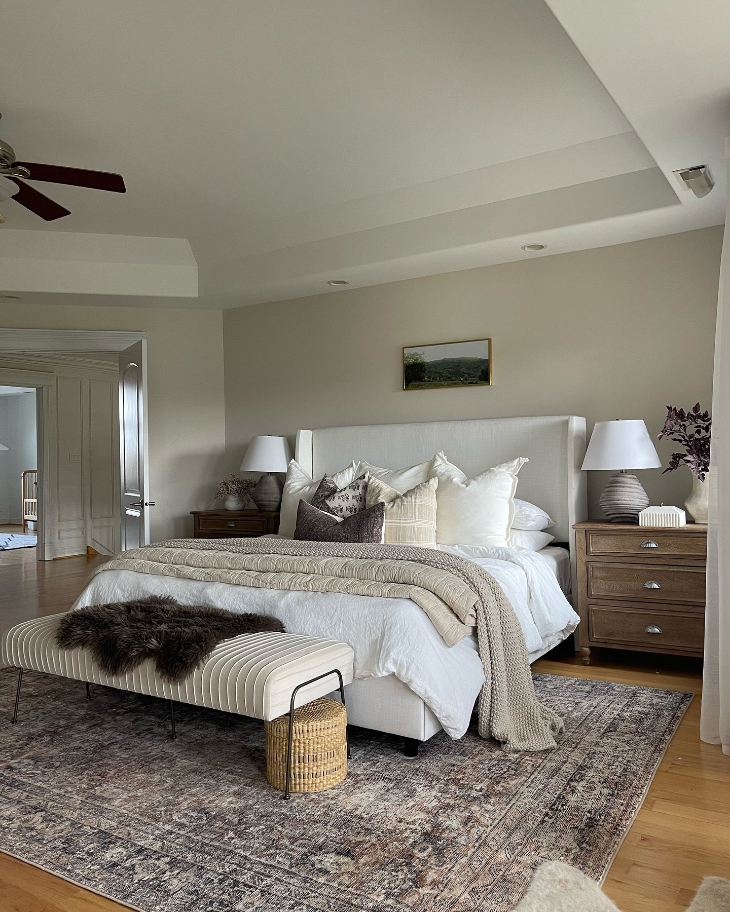 Spacious bedroom featuring a white upholstered bed, dark wood nightstands, a patterned rug, and a beige striped bench with a dark faux fur throw.
