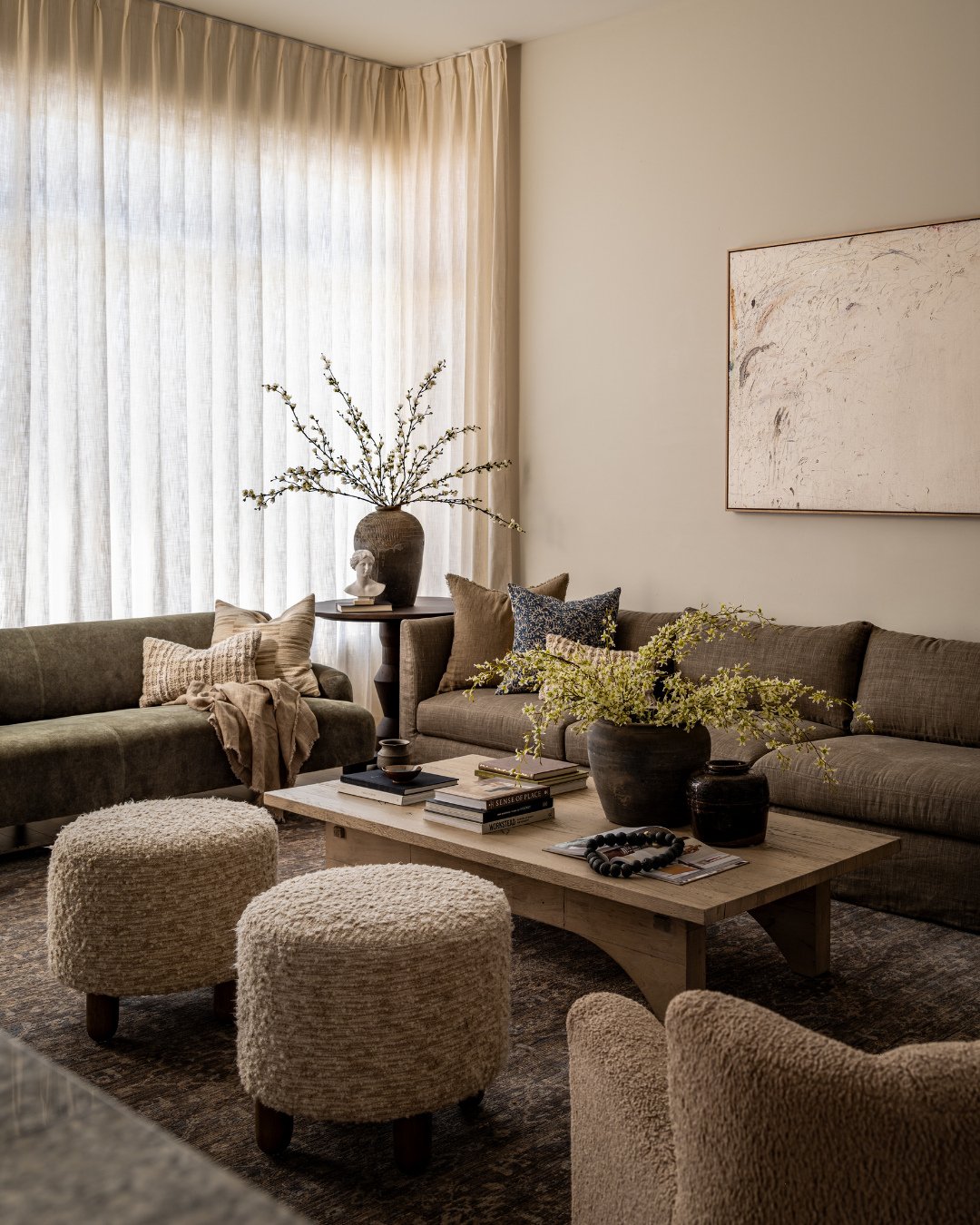 A cozy, modern living room featuring olive green sofas, a light wood coffee table, cream boucle ottomans, and sheer floor-to-ceiling curtains.