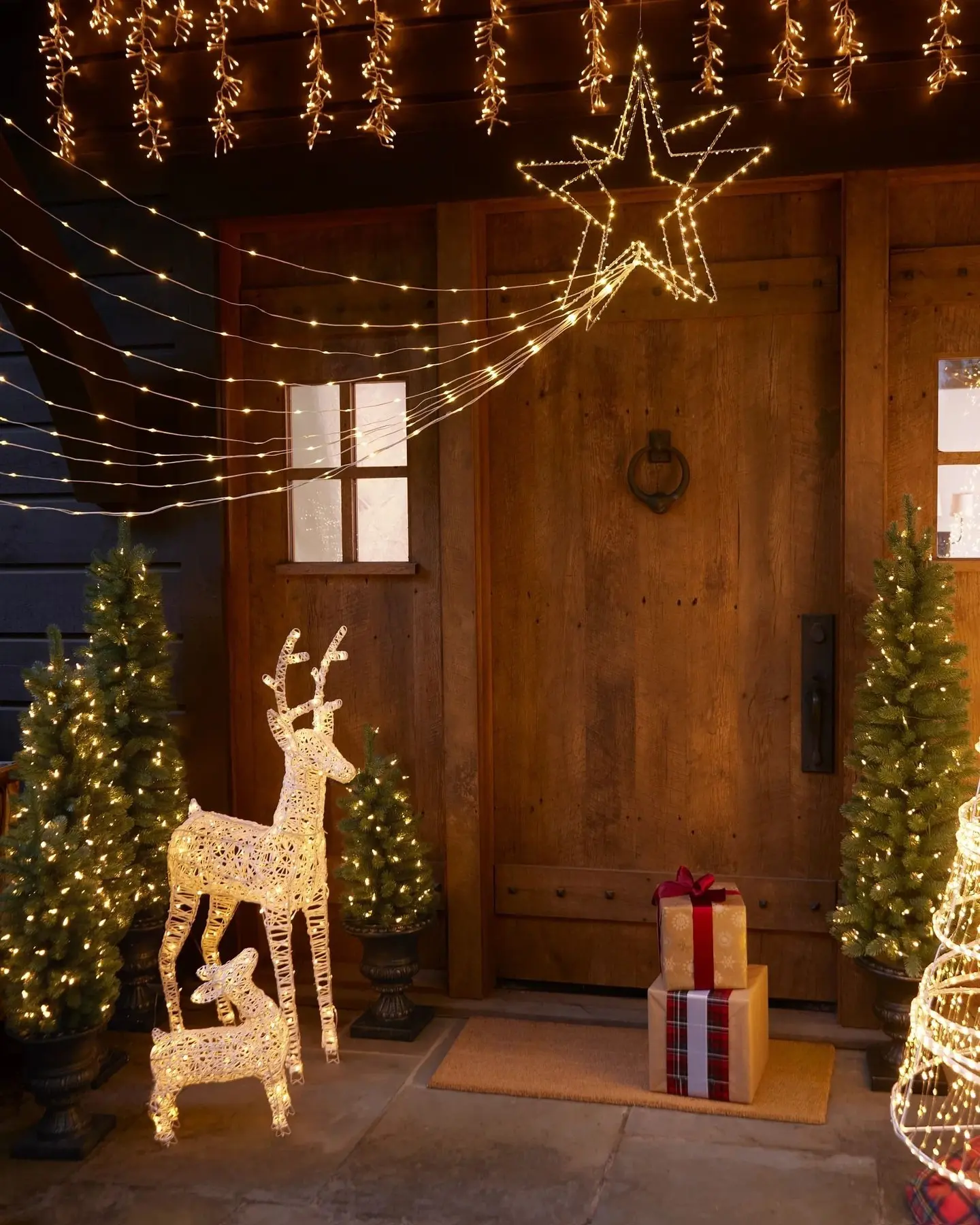 A rustic wooden door is decorated with a glowing star motif, flanked by a light-up reindeer family and multiple potted evergreen trees, all under a cascade of warm white icicle and draped lights.