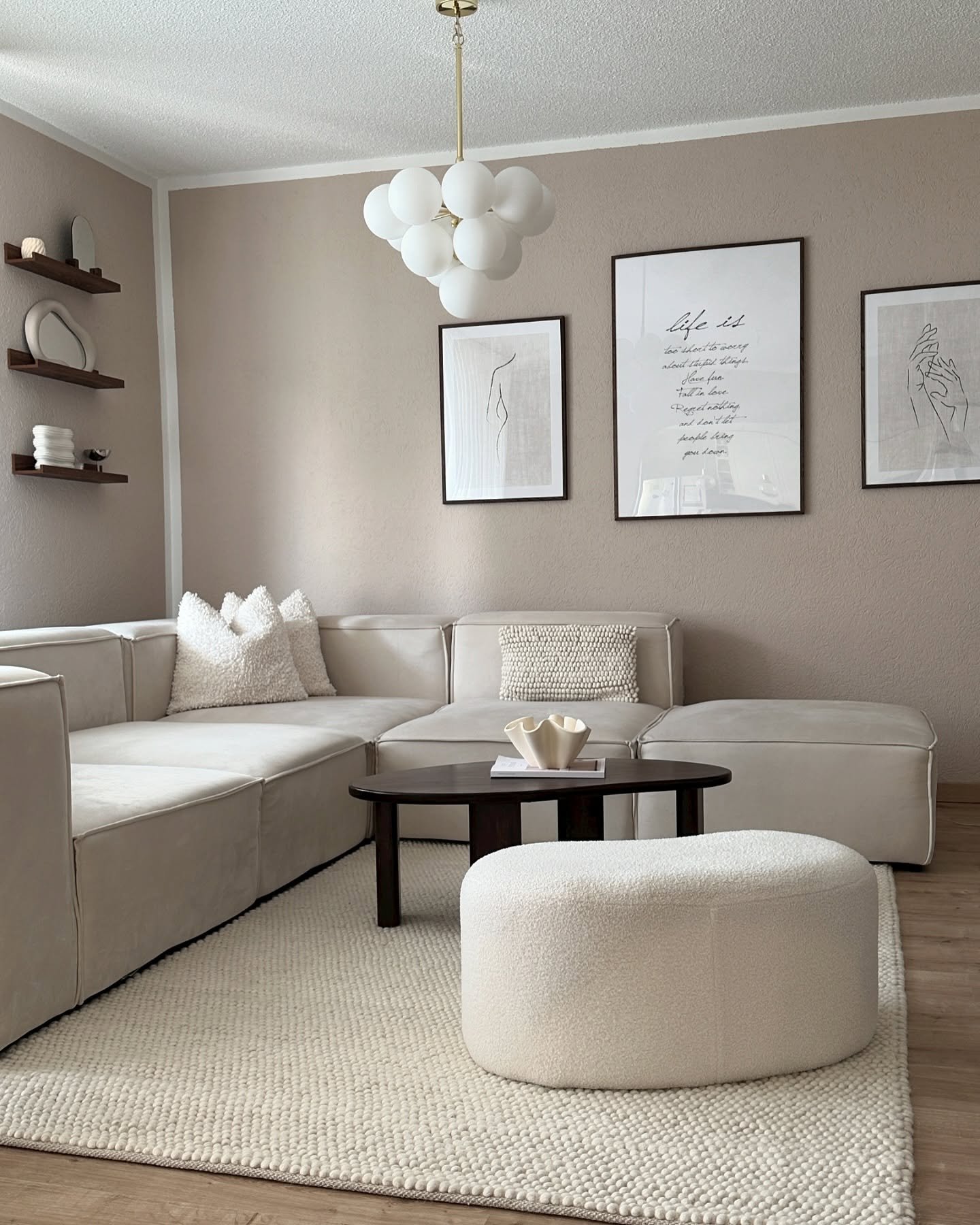 A contemporary beige living room featuring a deep taupe wall color, a square modular sofa, modern abstract art prints, and a spherical white chandelier.