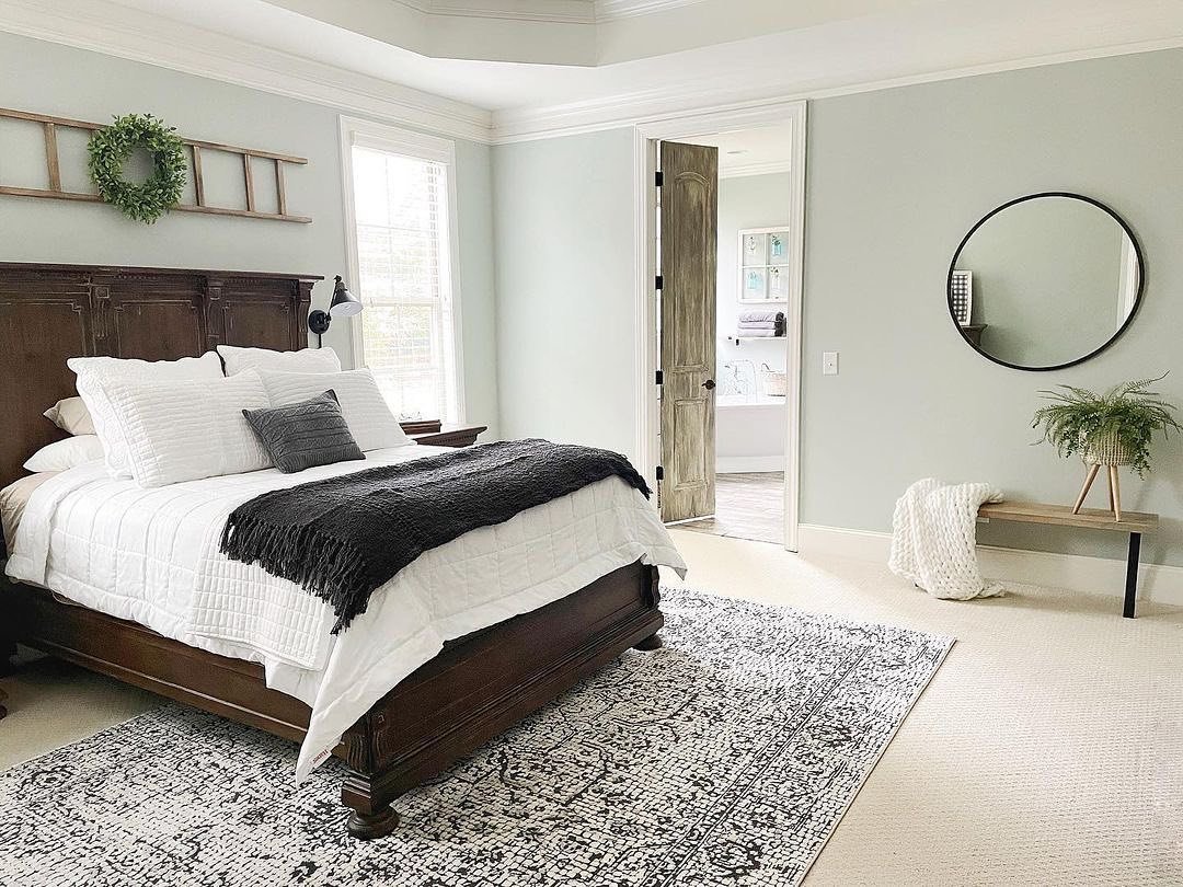 Bedroom painted in a light sage green with dark, traditional wood furniture, white bedding, and a black and white area rug.