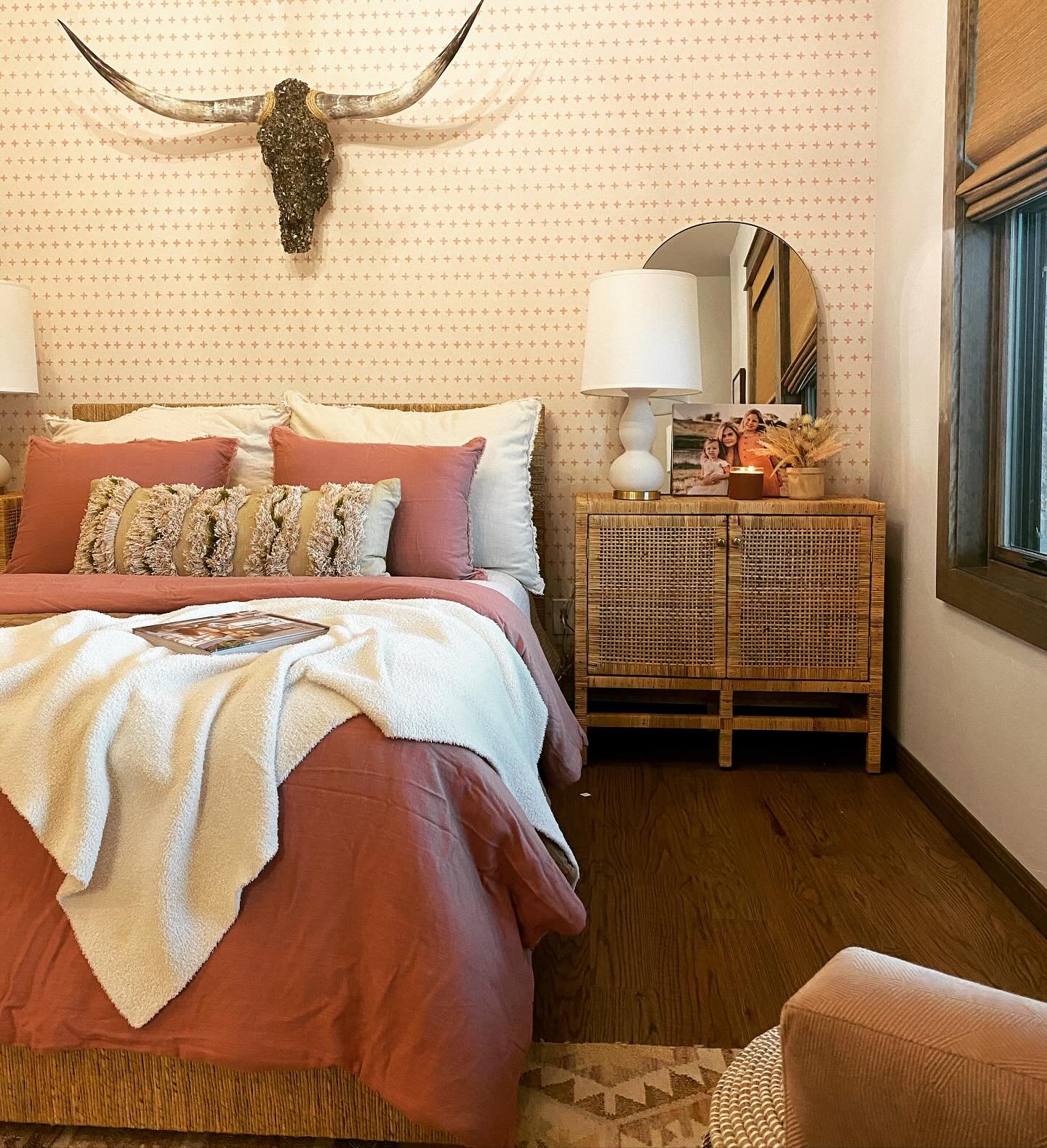 A soft Western-themed bedroom with pink bedding, cane nightstands, and a bronze longhorn skull mount on patterned wallpaper.