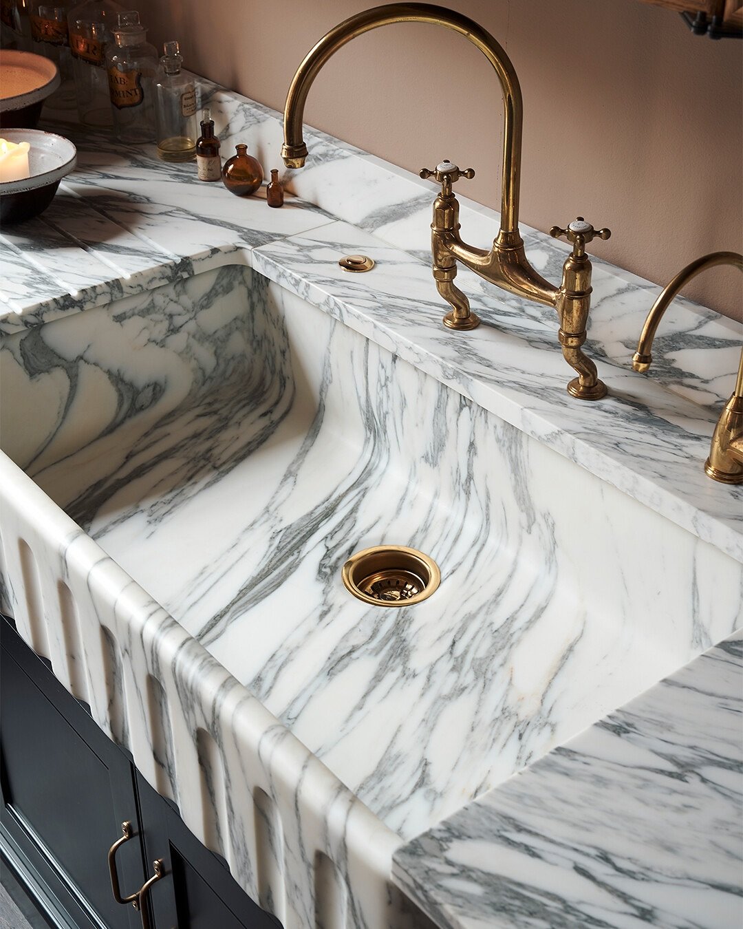 A luxurious single-bowl farmhouse sink carved entirely from white marble with gray veining, featuring a fluted apron-front and an antique brass bridge faucet.