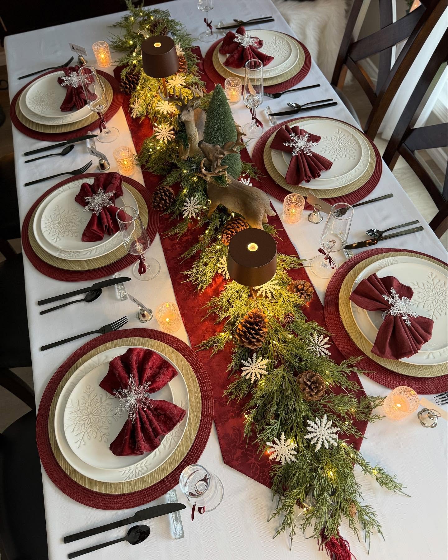 A Christmas dining table with a white cloth, set with a deep red runner, gold chargers, white snowflake-patterned plates, burgundy fan-folded napkins, and a centerpiece featuring pine branches, pinecones, tiny lights, and a decorative reindeer.
