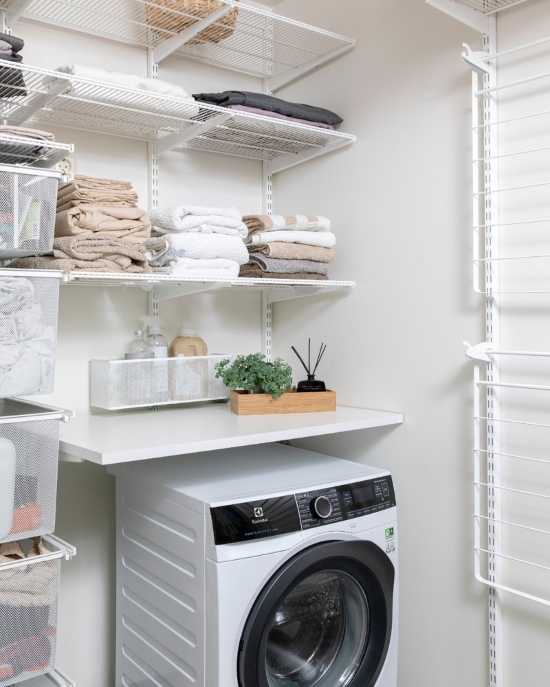 A small laundry closet utilizing a customizable white wire shelving system (like Elfa or similar). The system includes wire baskets, wire shelves, a solid shelf counter, and a vertical drying rack next to a washing machine.
