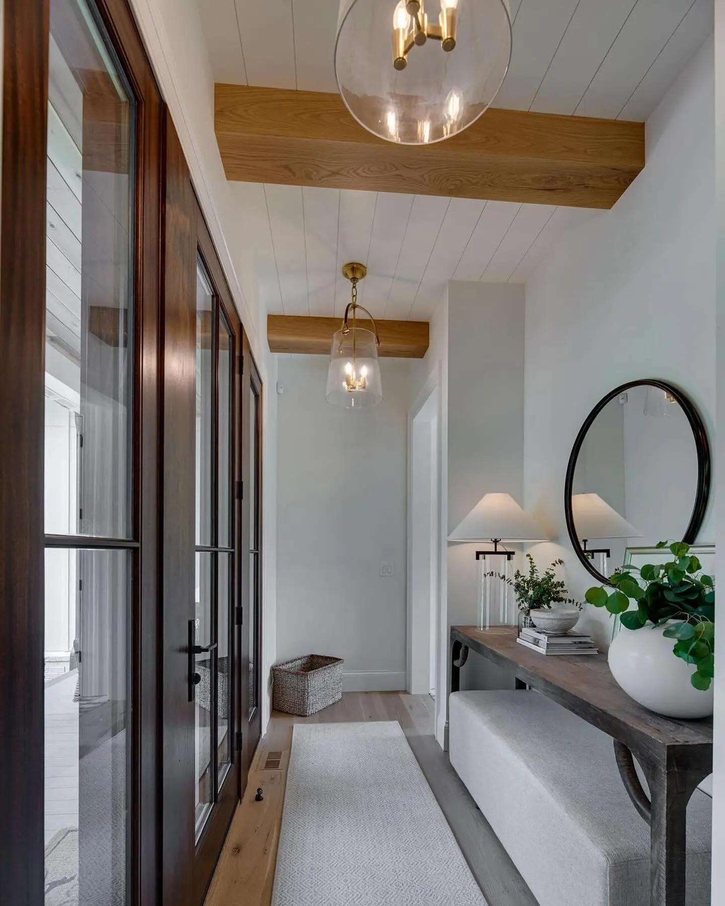 Small, bright entry hallway with dark wood and glass French doors, natural wood ceiling beams, shiplap ceiling, and two clear glass pendant lights.