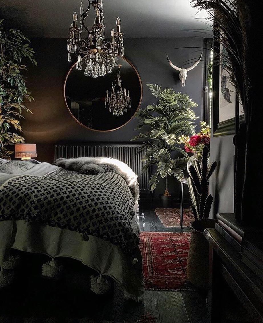 Dark, bohemian-style bedroom with matte black walls, a round copper-framed mirror, abundant indoor plants, a crystal chandelier, and a cow skull accent.