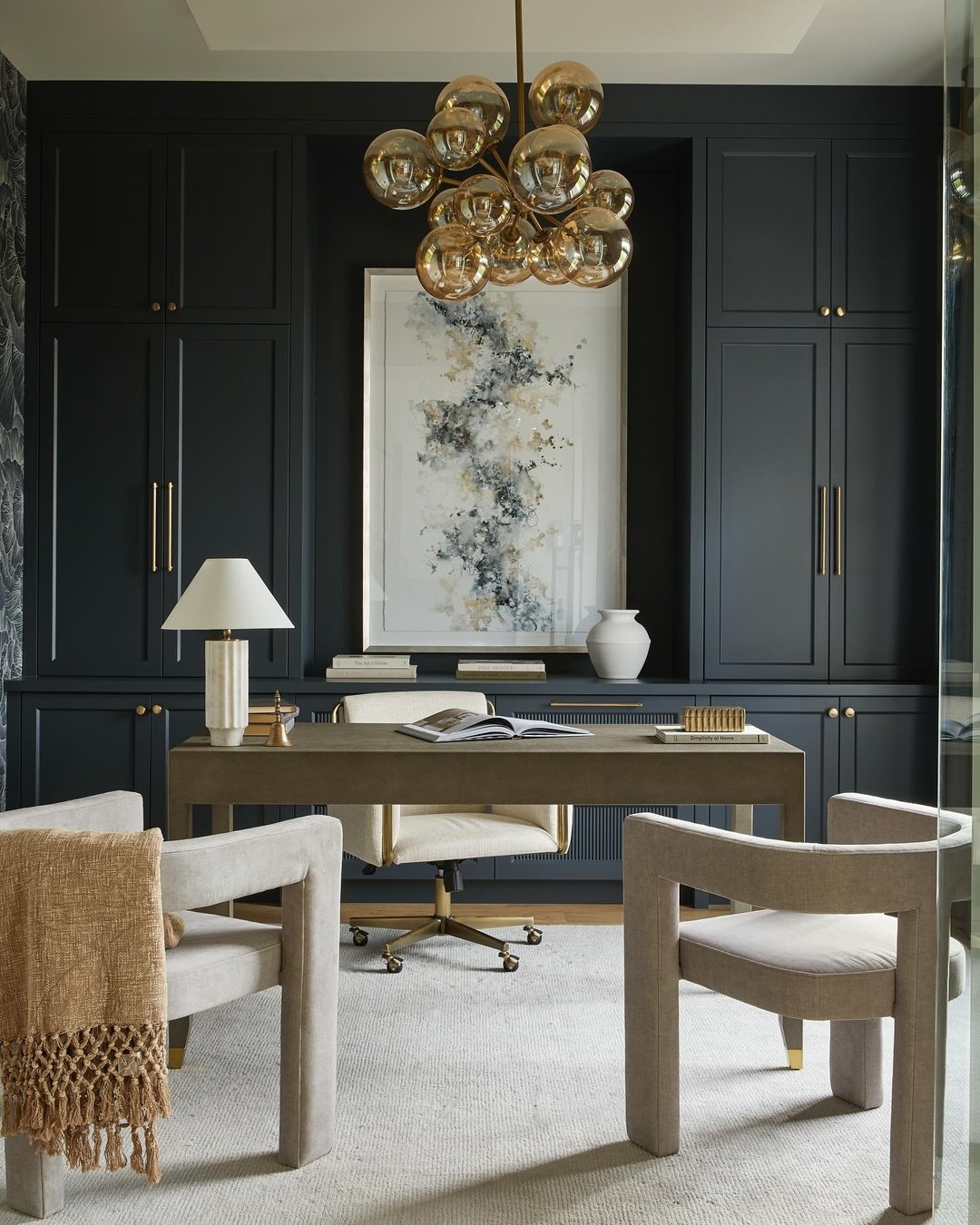 Luxurious home office with deep charcoal built-in cabinets, a large bubble-style gold chandelier, a light greige desk, and two modern beige guest chairs.