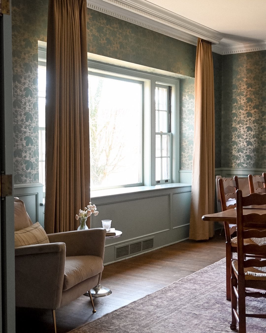 Rich camel-colored velvet curtain panels framing a large window in a room with blue-green paneling and damask wallpaper.