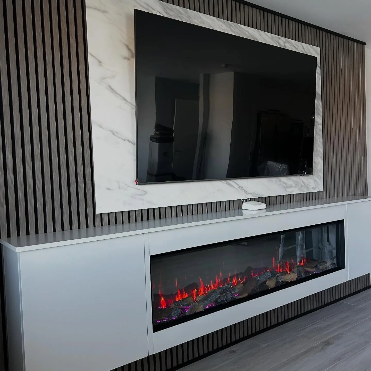 Ultra-modern living room featuring a dark wood vertical slat wall, an extra-wide floating media cabinet with an electric fireplace, and a TV framed by a white marble border.