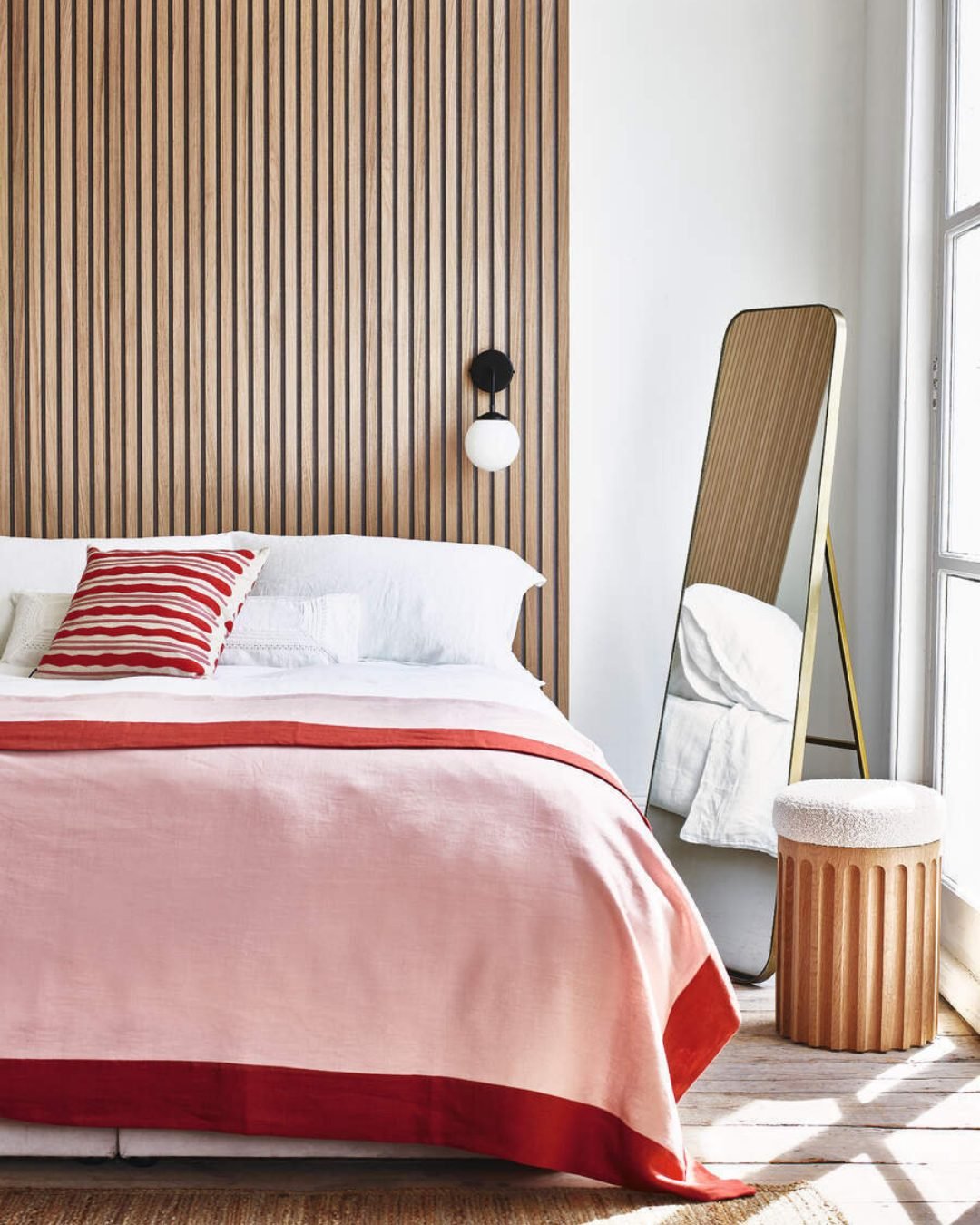 Full-height headboard wall covered in narrow, vertical wooden slats with dark backing, illuminated by a modern black and white globe sconce beside a bed with pink and red linen.