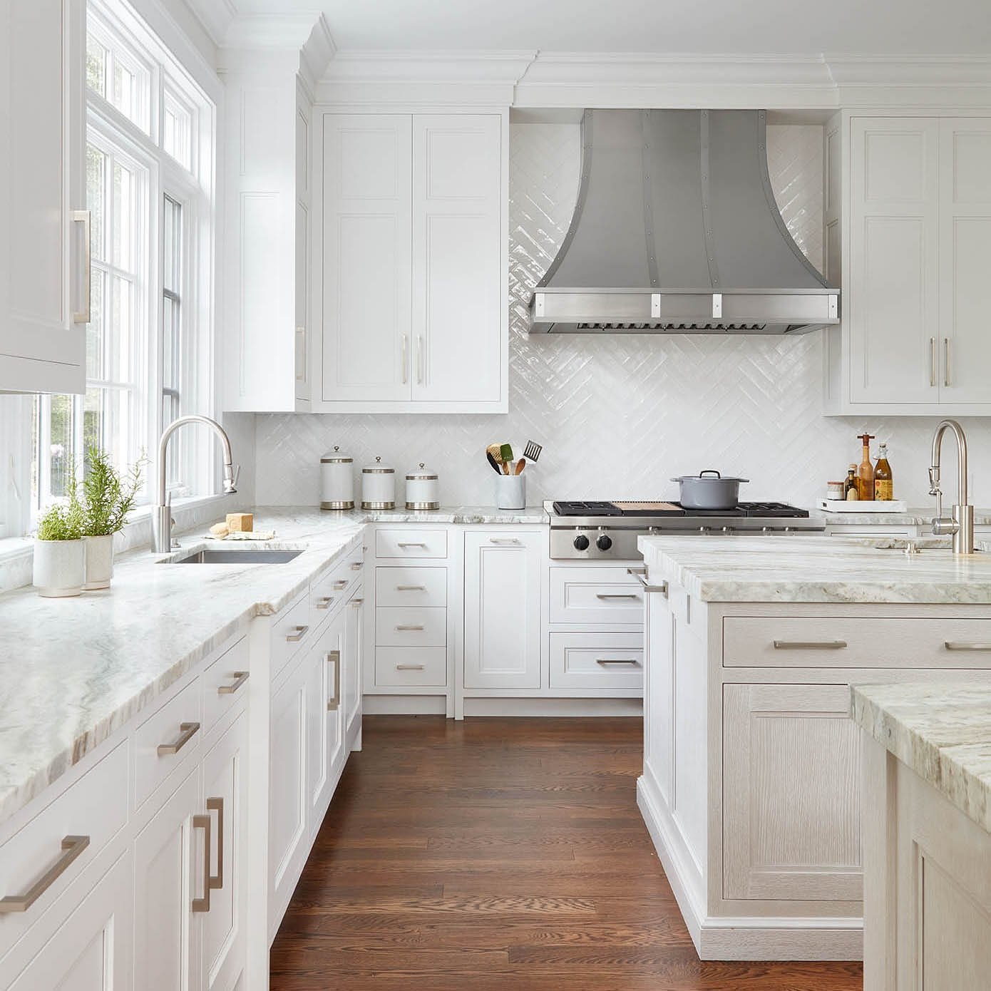 Bright white kitchen with white shaker cabinets, gray and white marble countertops, a glossy white herringbone tile backsplash, and a large stainless steel range hood.