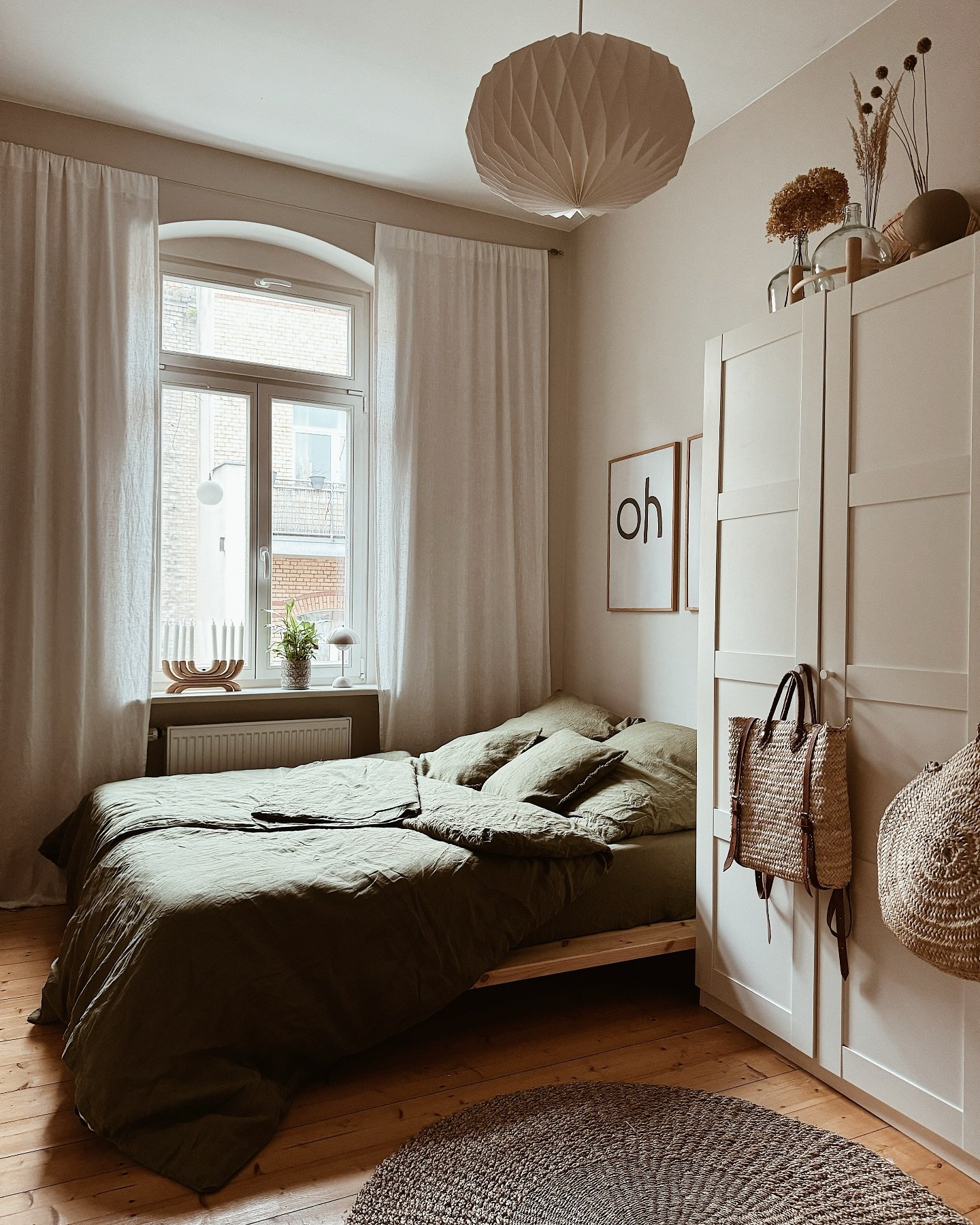 A Scandinavian-style bedroom with warm wood floors, white walls, and a low-profile bed dressed in wrinkled olive green linen bedding.