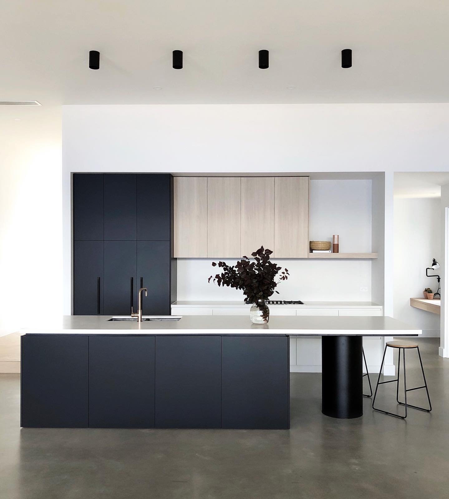 Contemporary kitchen featuring dark charcoal cabinetry, light wood wall units, a large island with a column base, and a polished concrete floor.