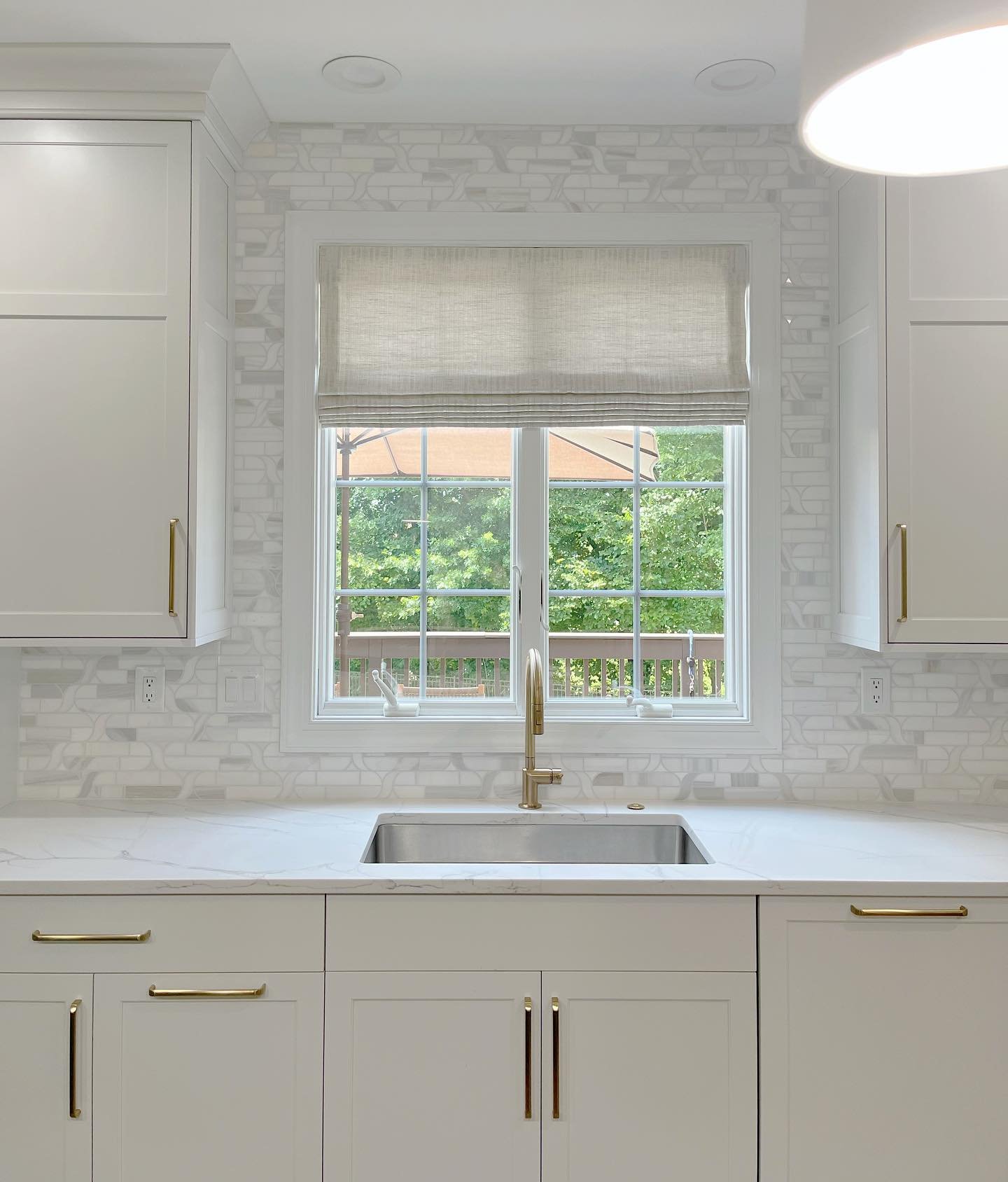 White transitional kitchen with light cabinets and gold hardware. A white grid window is centered over a stainless steel sink and surrounded by a mosaic marble tile backsplash.