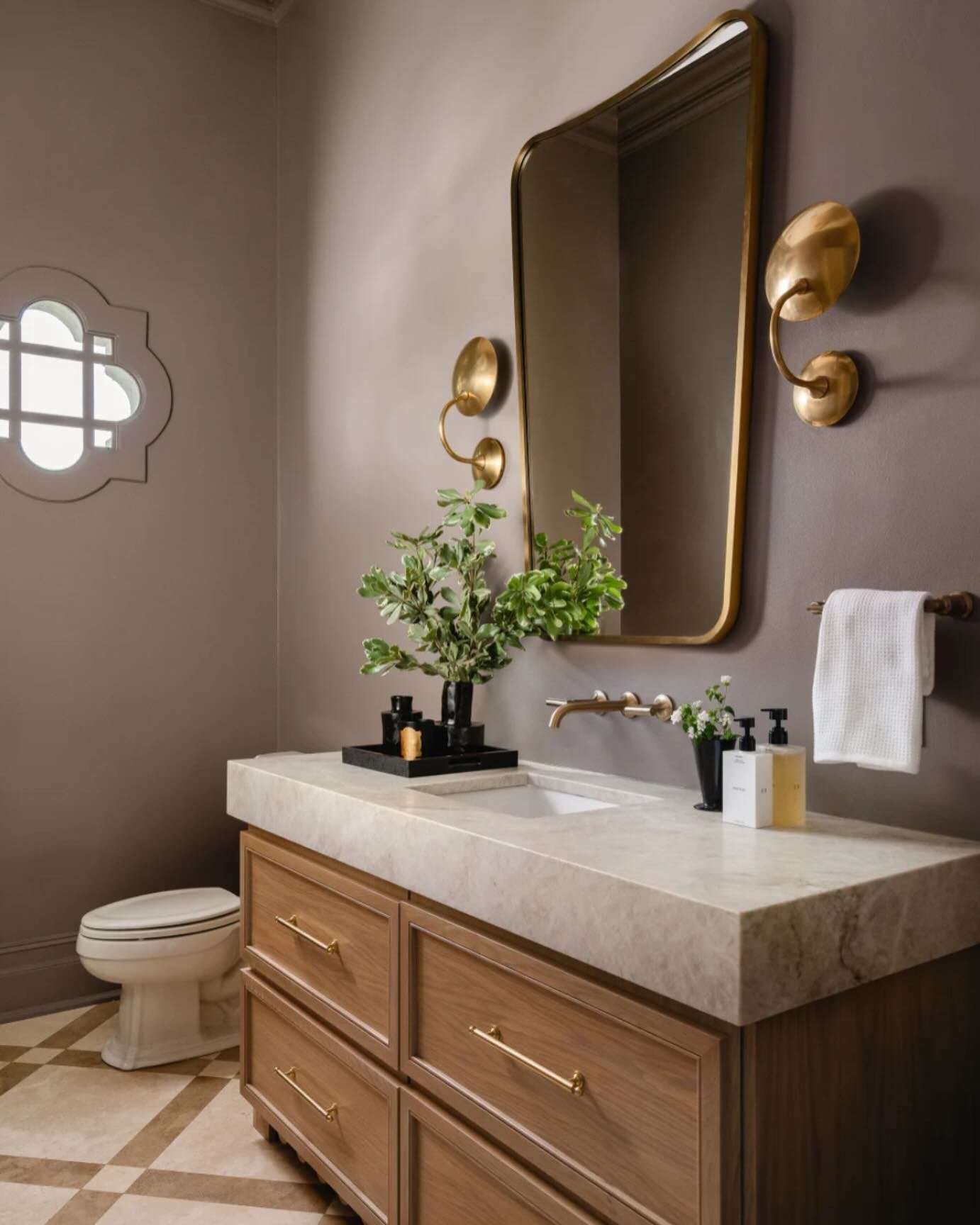 Taupe-colored powder room with a large wooden vanity, marble countertop, and a unique architectural ogee-shaped window.