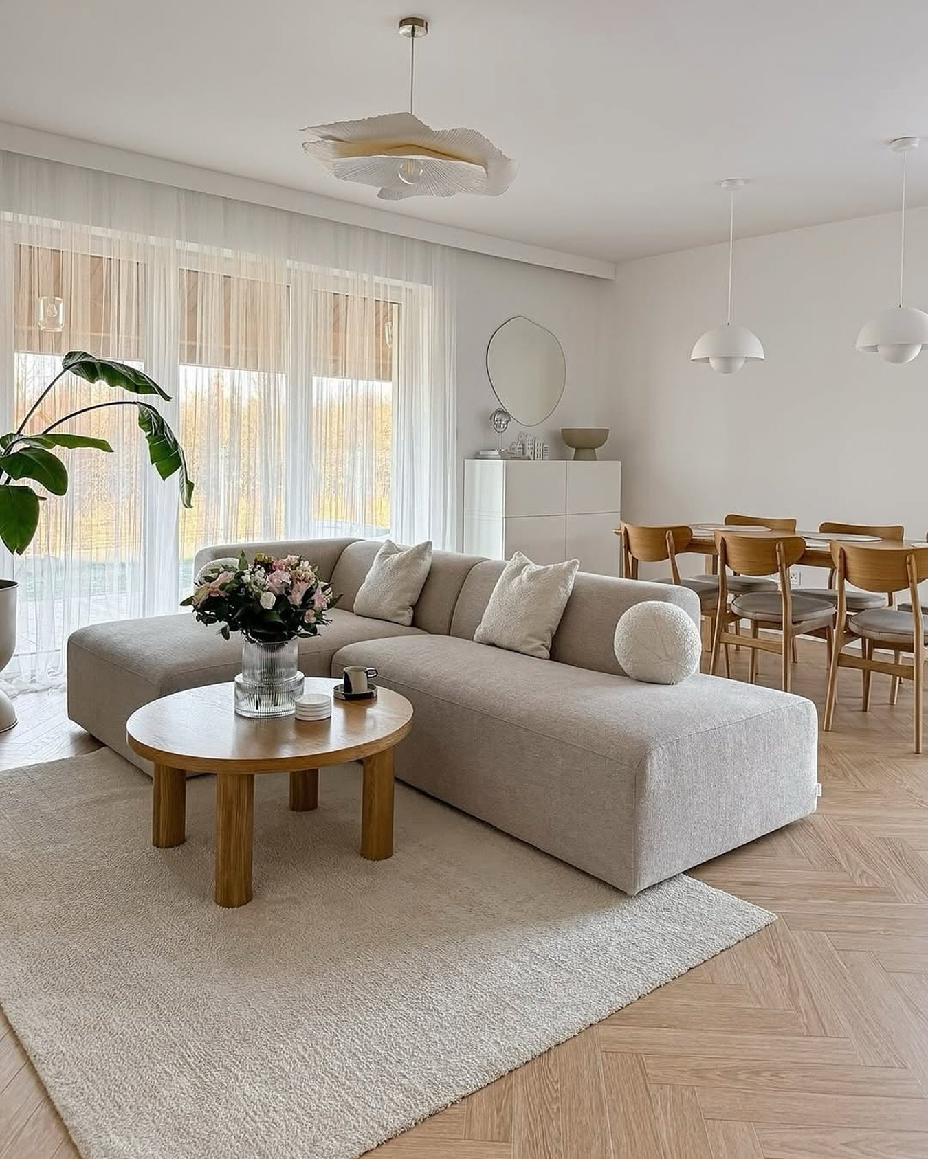 An open-concept living and dining area with a low, light beige sectional, a round wood coffee table, and light wood flooring laid in a herringbone pattern.