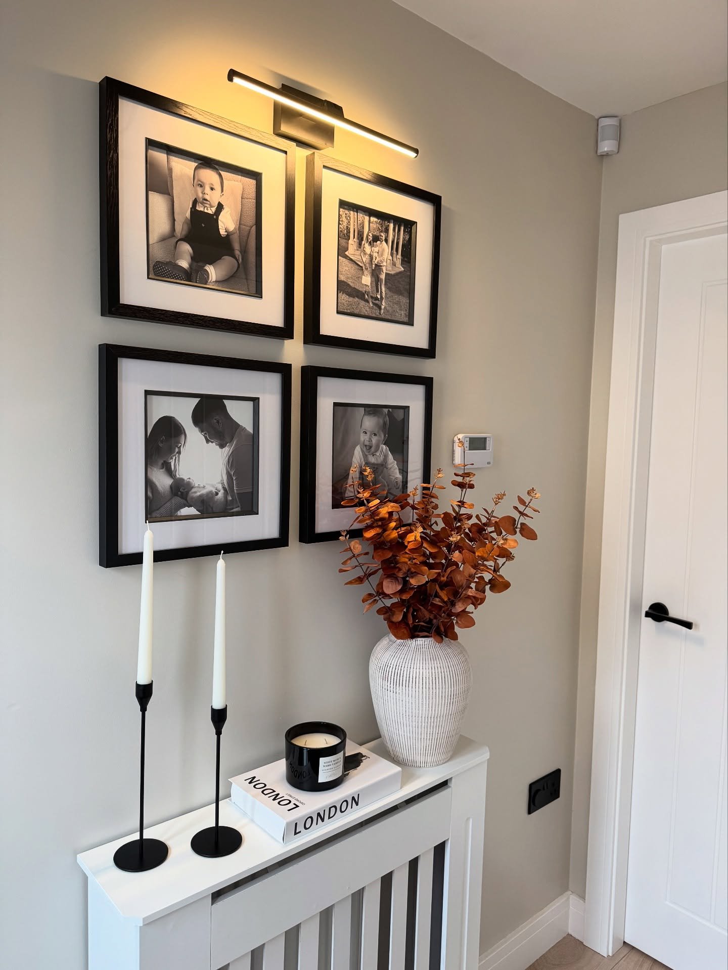 A white slatted radiator cover in a light taupe hallway topped with a gallery wall of four black and white photos, white candles, and a white vase with orange foliage.