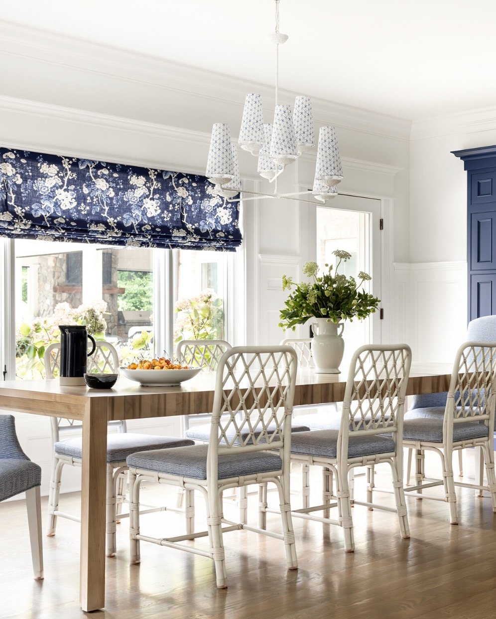 A bright dining room with a light wood table, white faux bamboo dining chairs with lattice backs, and a navy floral patterned Roman shade and chandelier shades.