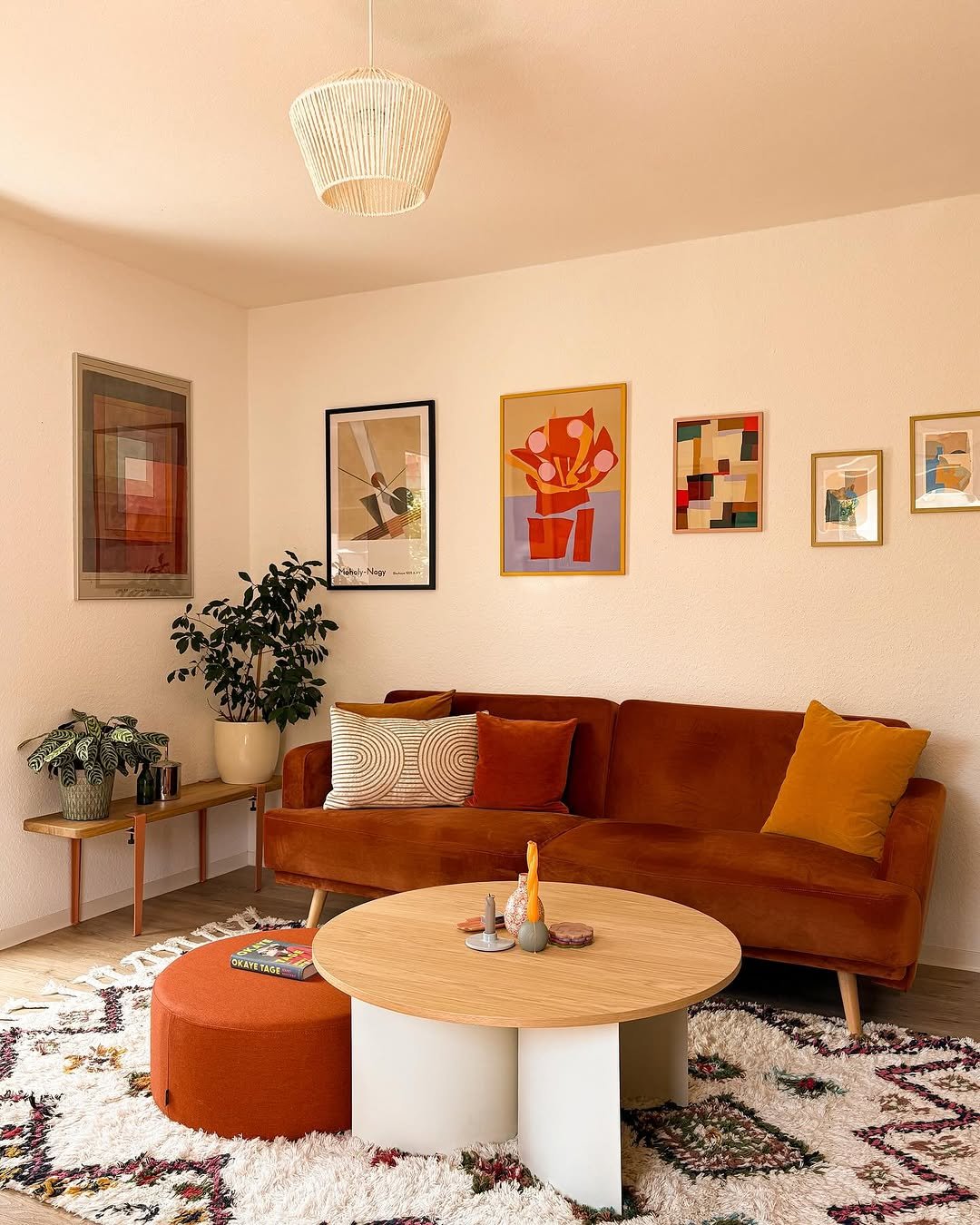 A small, eclectic living room featuring an orange velvet sofa, a gallery wall of colorful abstract art, a shaggy Berber rug, and an orange upholstered ottoman.