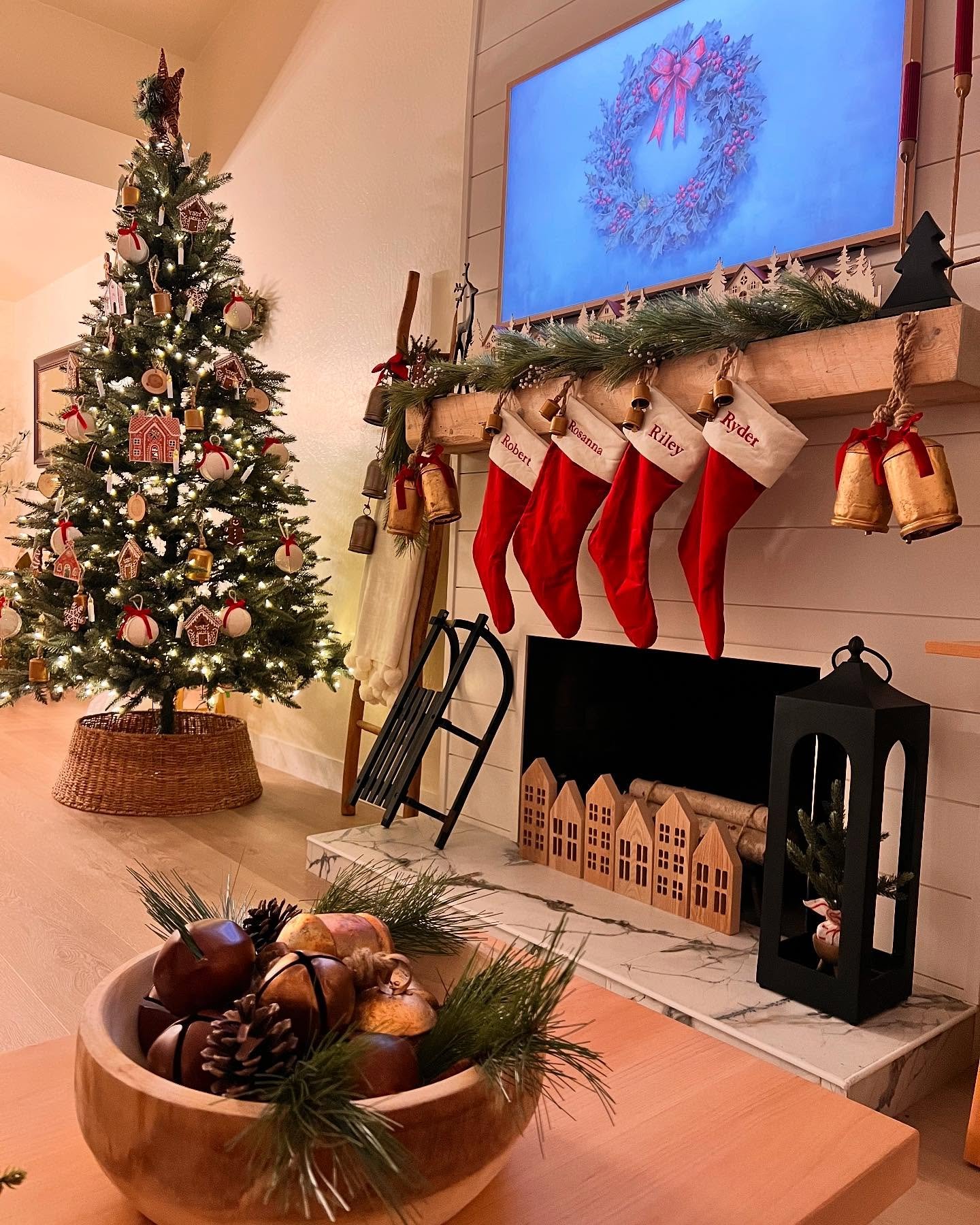 Rustic wood mantel with four personalized red felt stockings, a simple pine garland, and large vintage-style brass bells hanging on ropes.