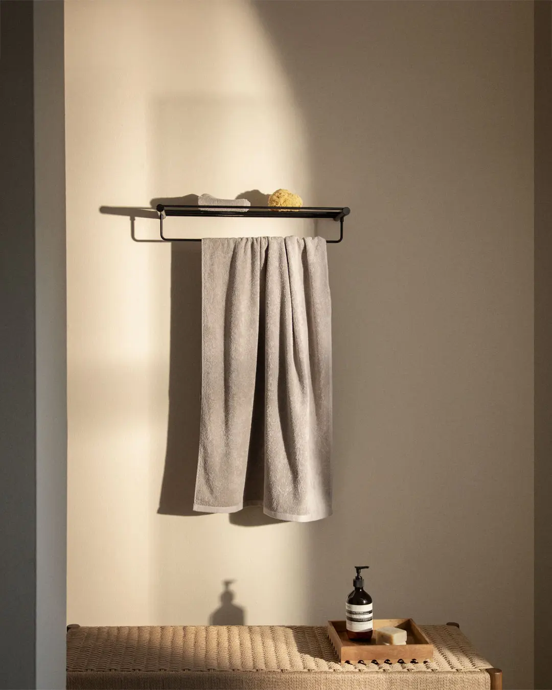 Wall-mounted matte black hotel rack with a flat shelf on top and a horizontal bar below, holding a light grey bath towel and a natural sponge.