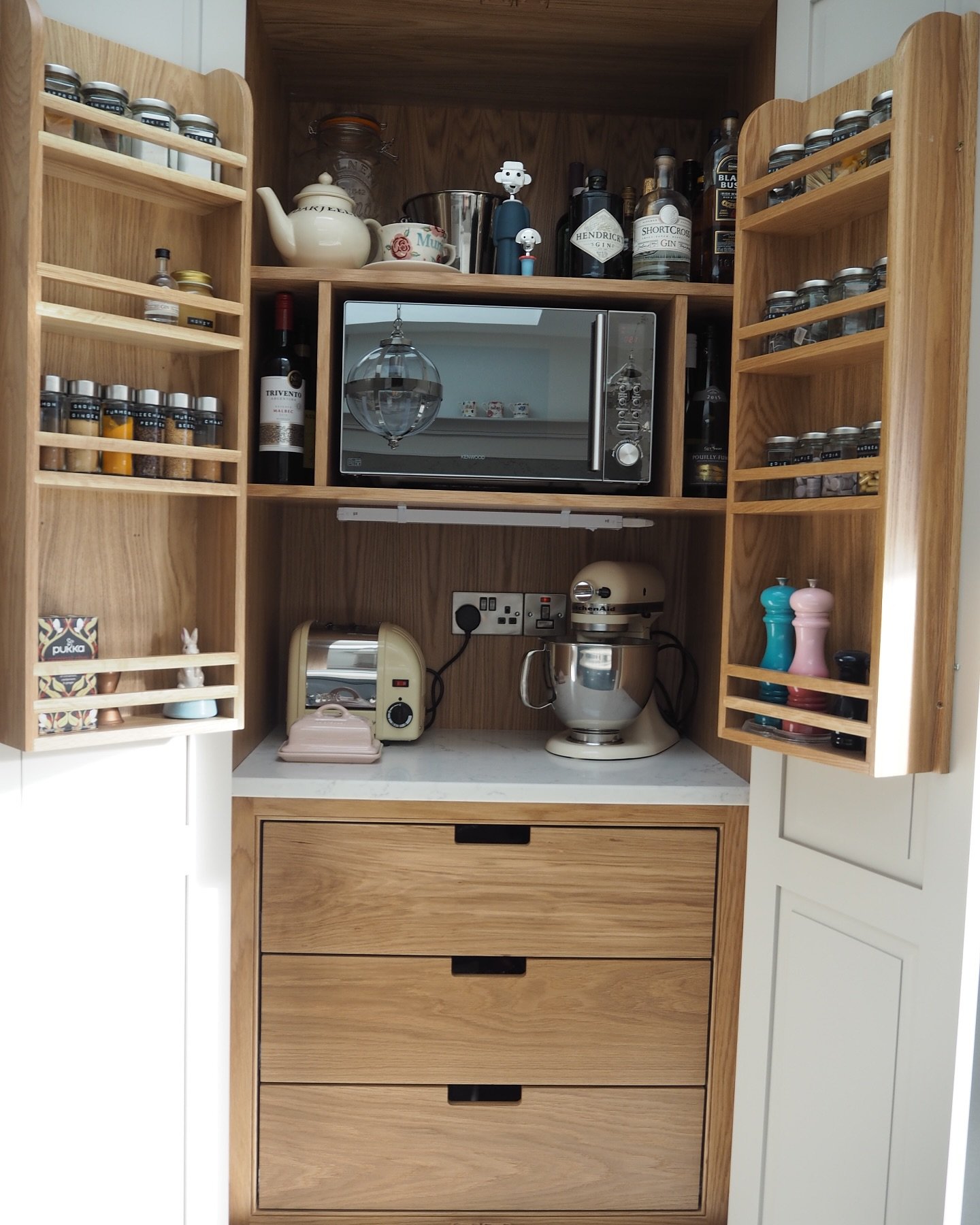 Tall wooden kitchen cabinet open to show a counter area housing a stand mixer and toaster, with spice racks mounted on the inside of both doors.