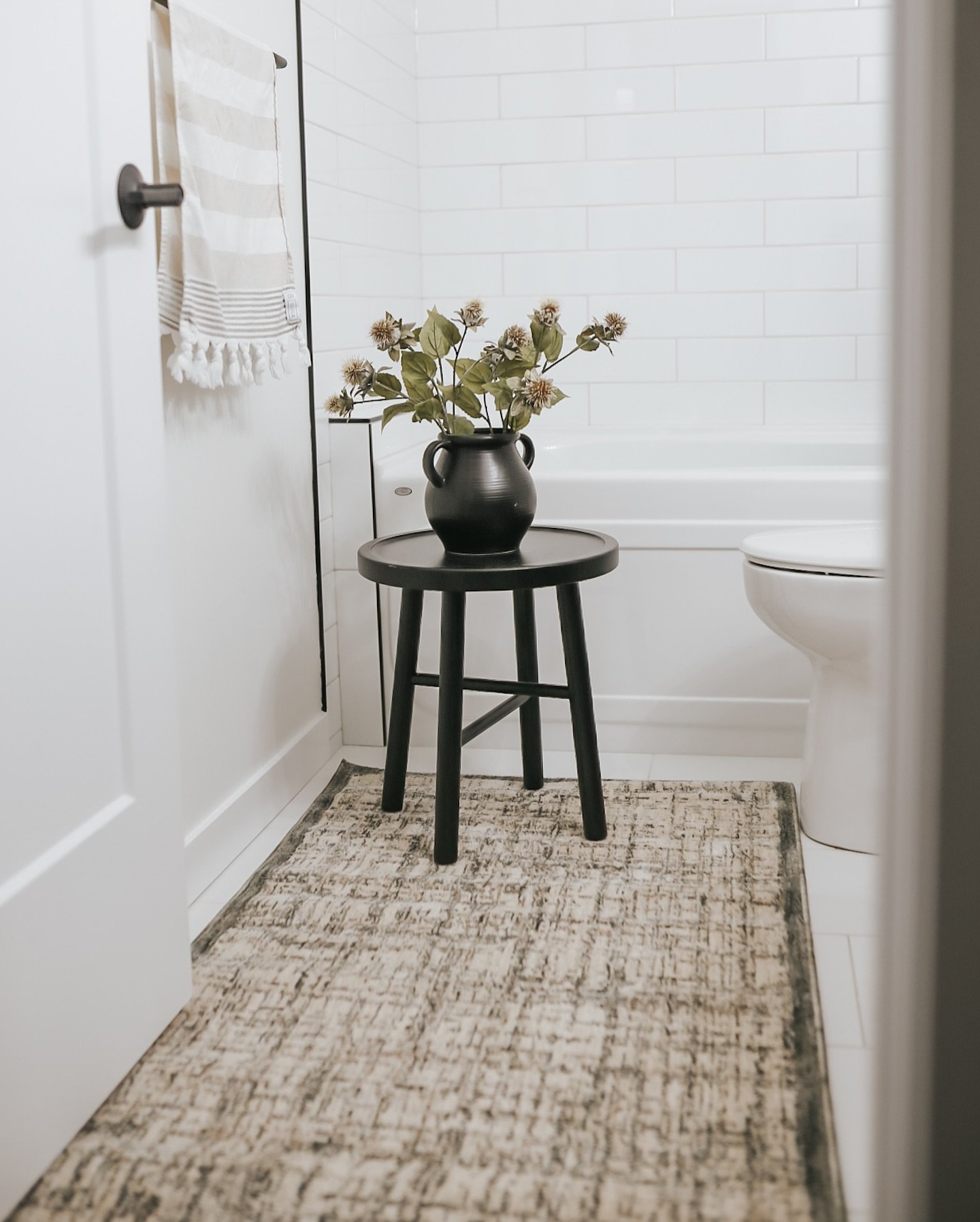 A clean, all-white bathroom with subway tile walls, a toilet, a small black side table with a vase of flowers, and a rectangular abstract patterned rug in neutral beige and gray tones.